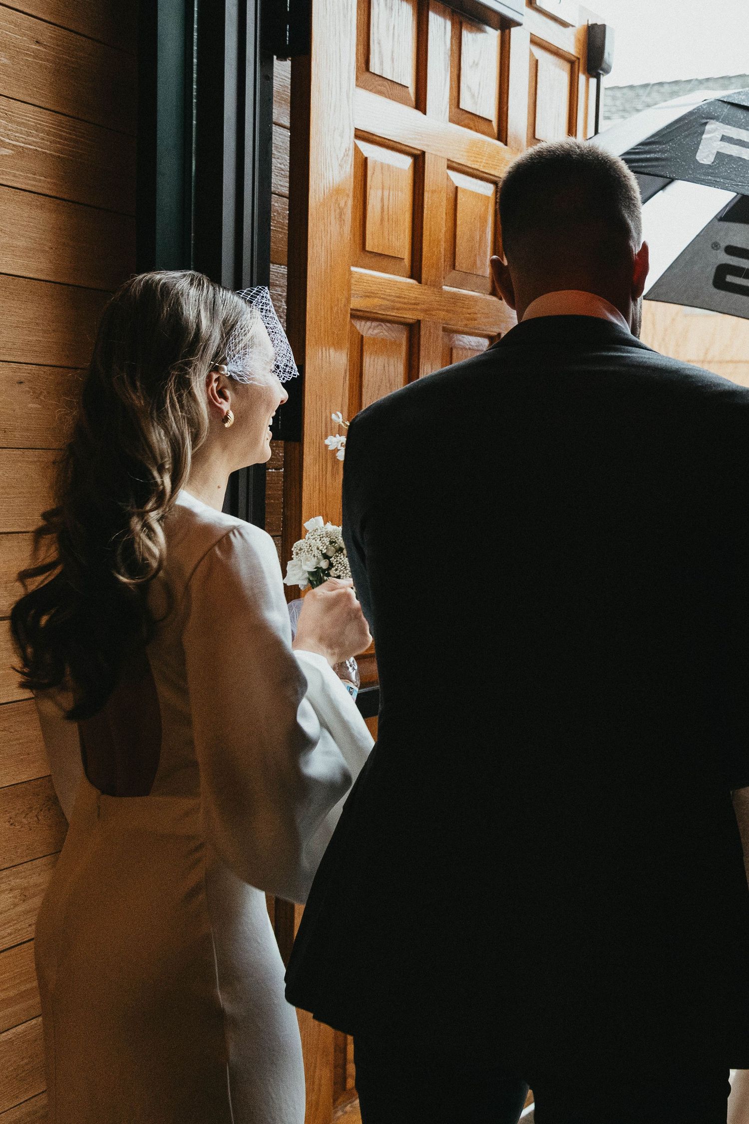 Bride in elegant white gown and veil stands with groom at wooden church doors before ceremony.