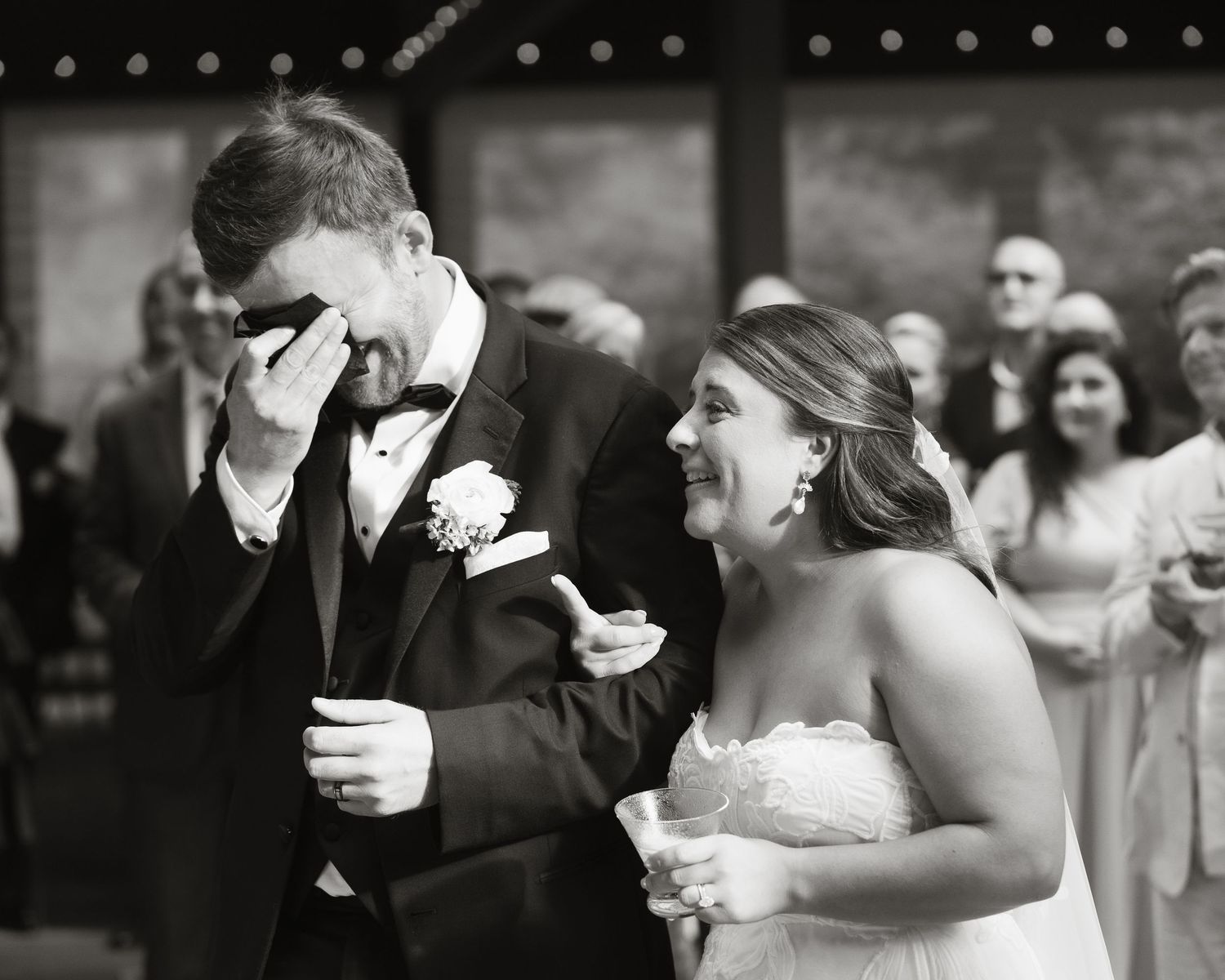 Black and white photo of emotional groom wiping tears while laughing bride holds his arm at wedding ceremony.