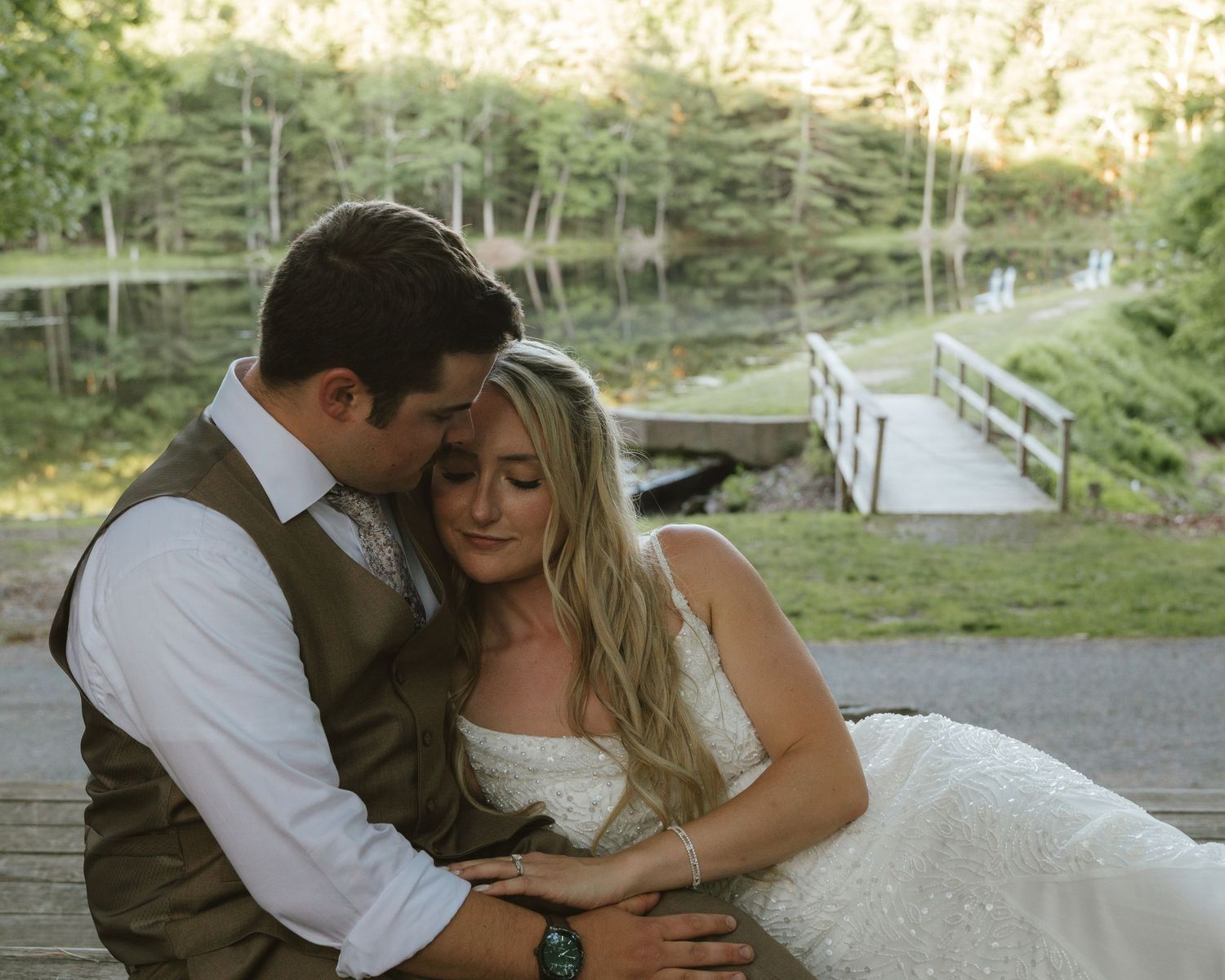 A bride and groom share a tender moment seated outdoors near a scenic pond and wooden bridge on their wedding day.