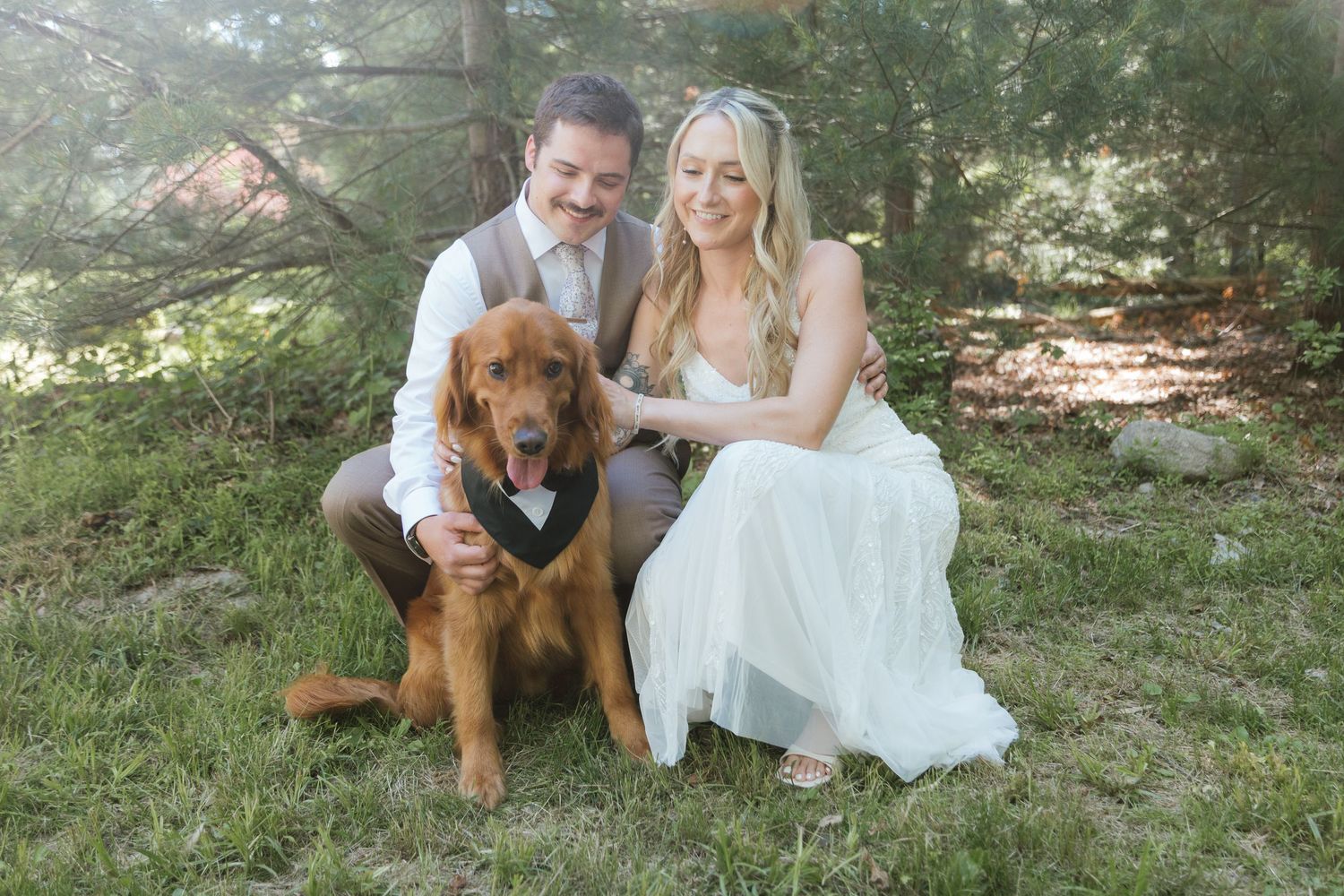 Bride and groom pose with their golden retriever dog wearing a bow tie outdoors in a forest wedding photo.