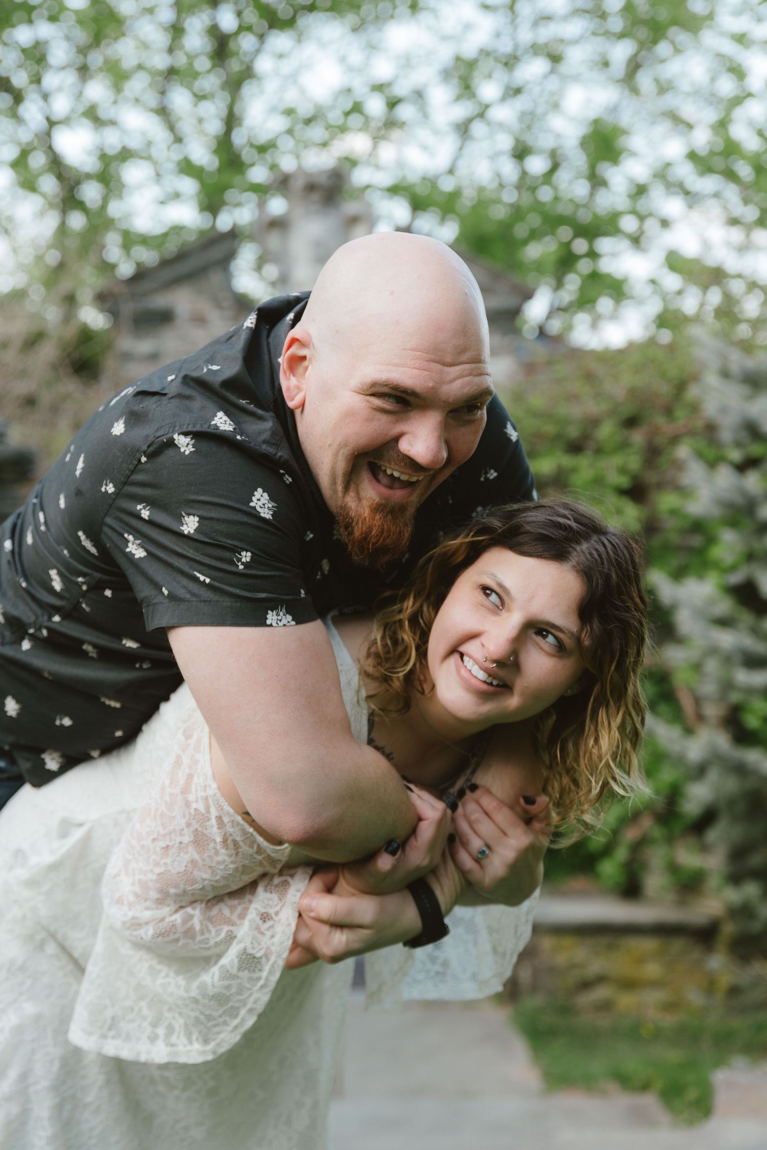 Laughing couple embracing outdoors, bald bearded man hugging woman in white lace dress in a garden setting.