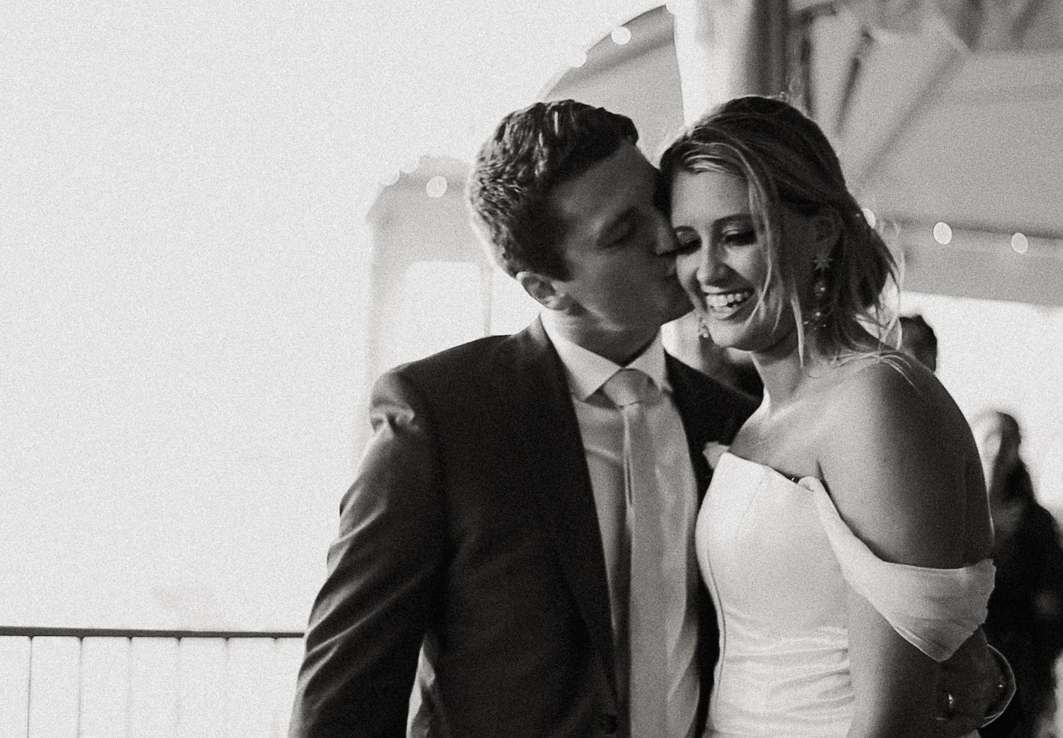 Black and white candid photography of a groom kissing a laughing bride's cheek at a wedding reception in the Poconos.
