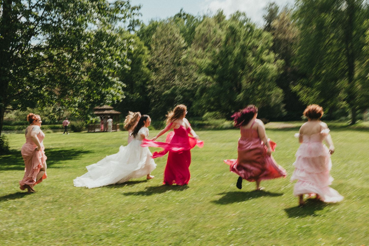 Bridesmaids in colorful dresses running across green lawn with bride in white wedding dress at outdoor celebration.
