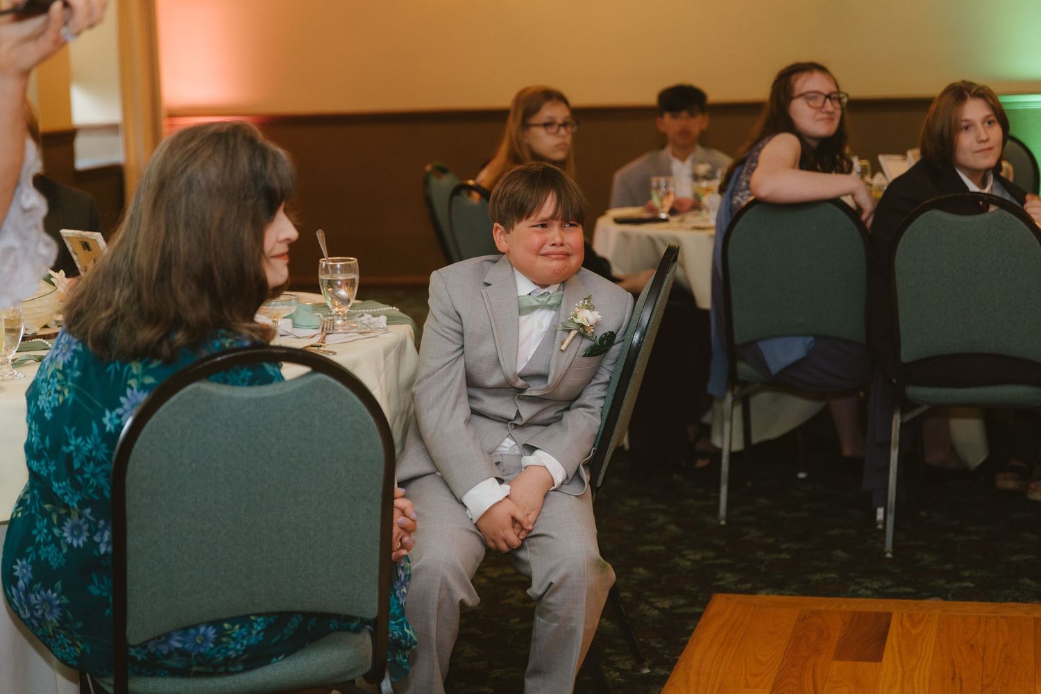 Young boy in gray suit smiling at wedding reception banquet hall with guests seated at round tables.
