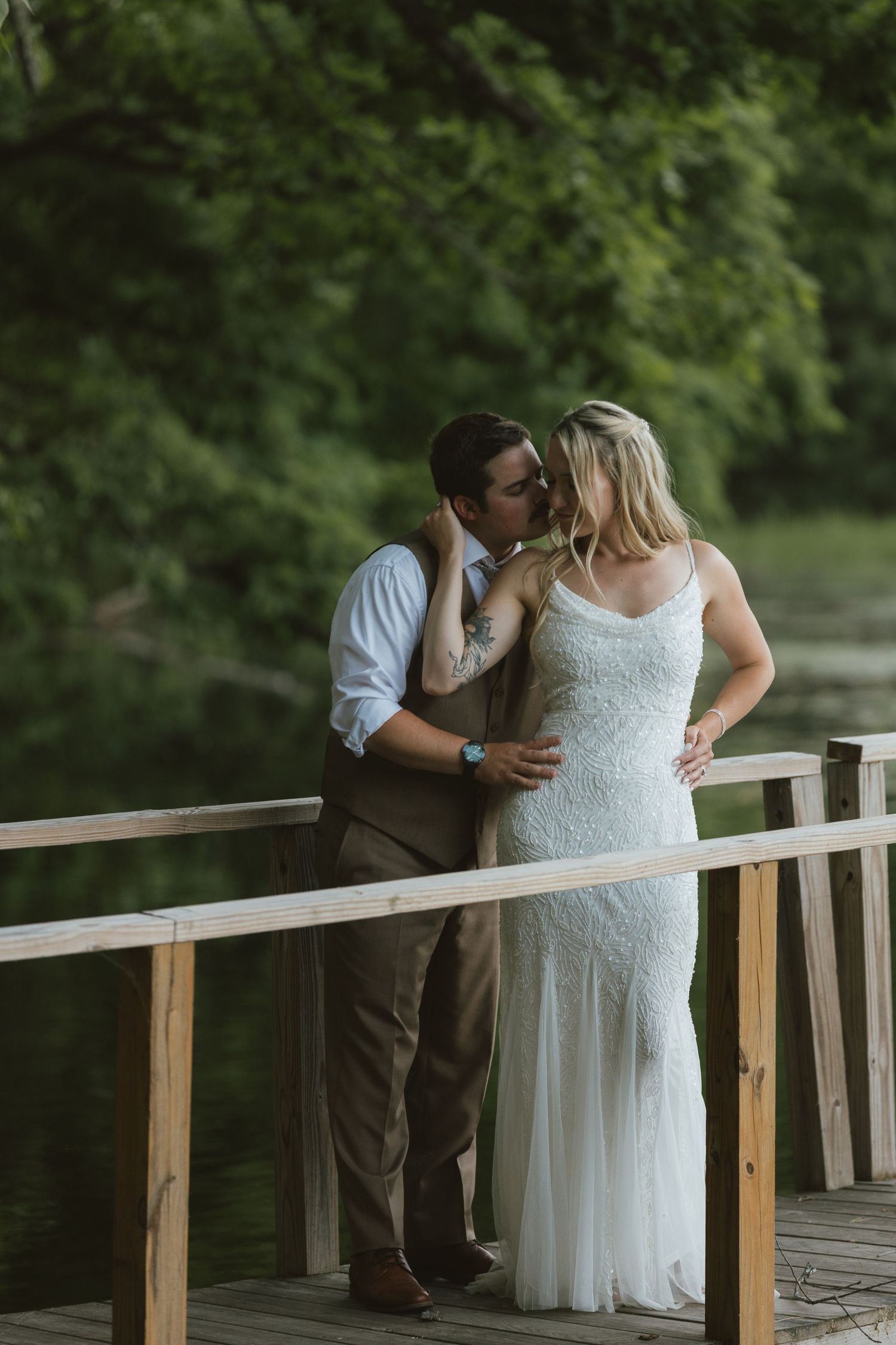 Bride and groom share a romantic kiss on a wooden bridge surrounded by lush green trees near a serene lake.
