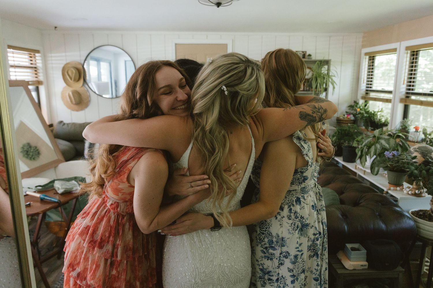 Three women in floral dresses share a warm group hug inside a cozy, plant-filled living room.
