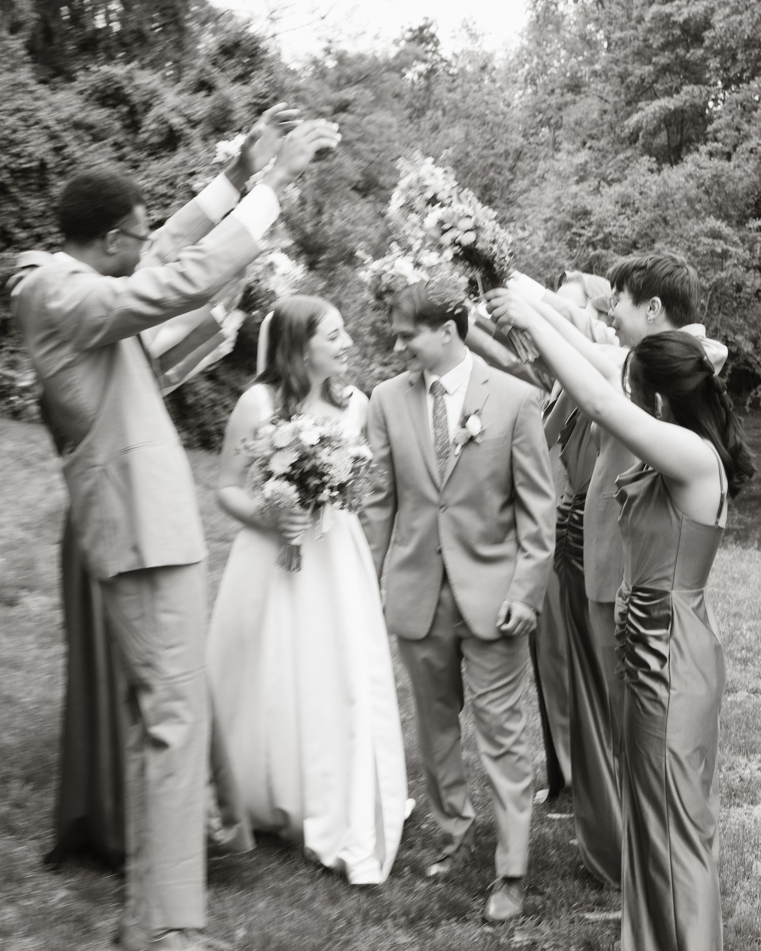 Black and white photo of wedding party raising bouquets over smiling bride and groom.