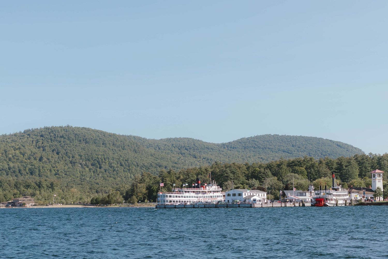 A white steamboat docked near a forested hillside on a calm blue lake under clear skies.