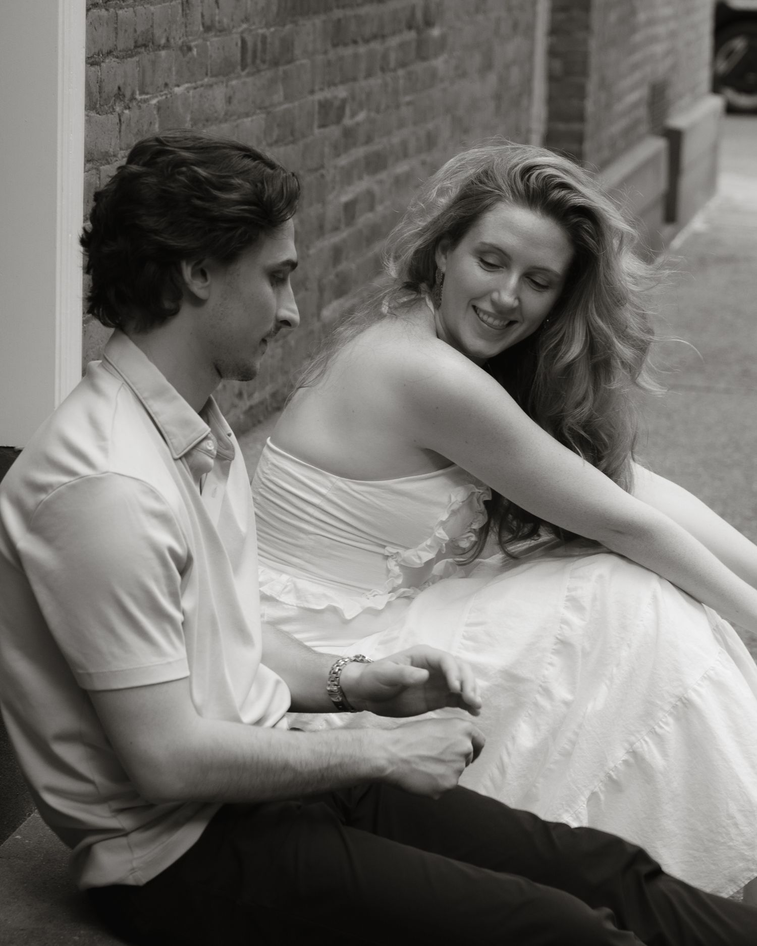 Black and white photo of a smiling couple sitting together holding hands on an urban sidewalk.