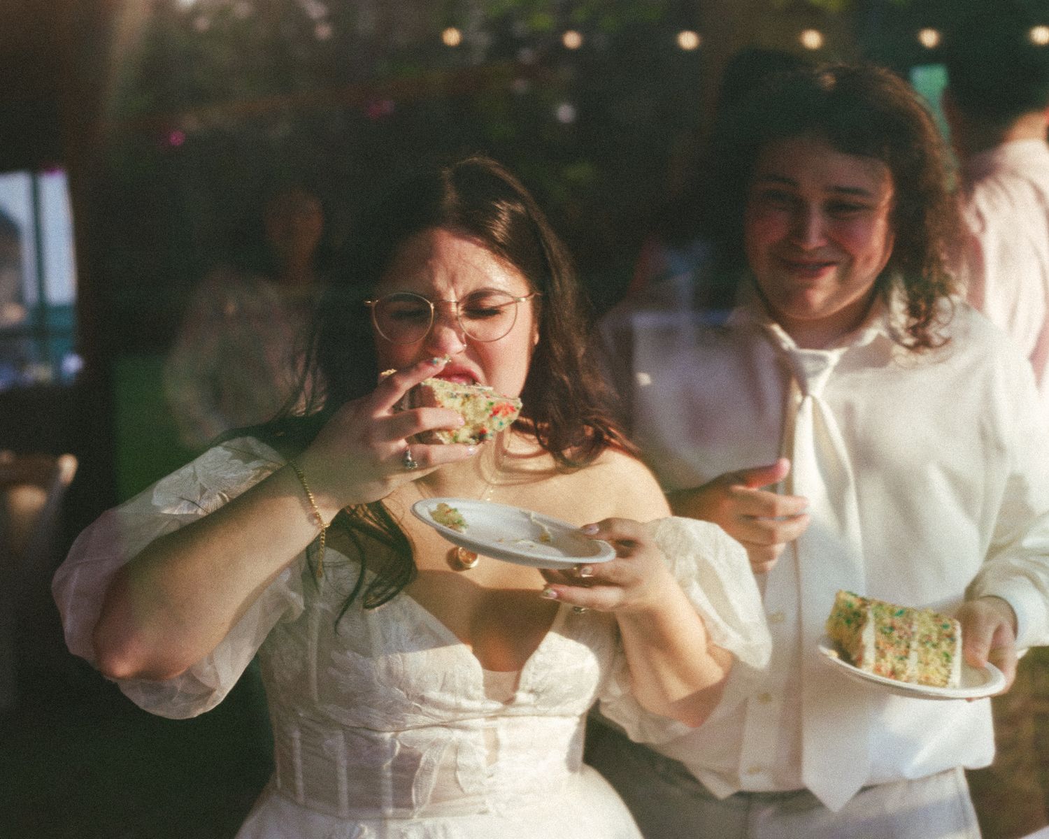 Bride enthusiastically eating wedding cake while guest in white looks on smiling at reception.