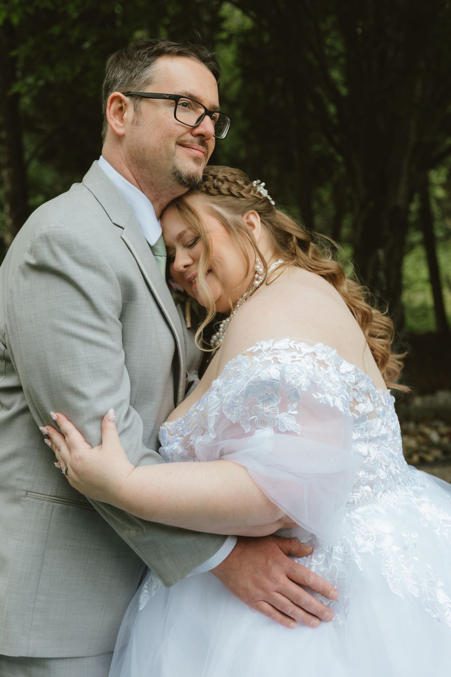 A bride in a white lace gown rests her head on her groom's chest as he holds her in a forest setting.