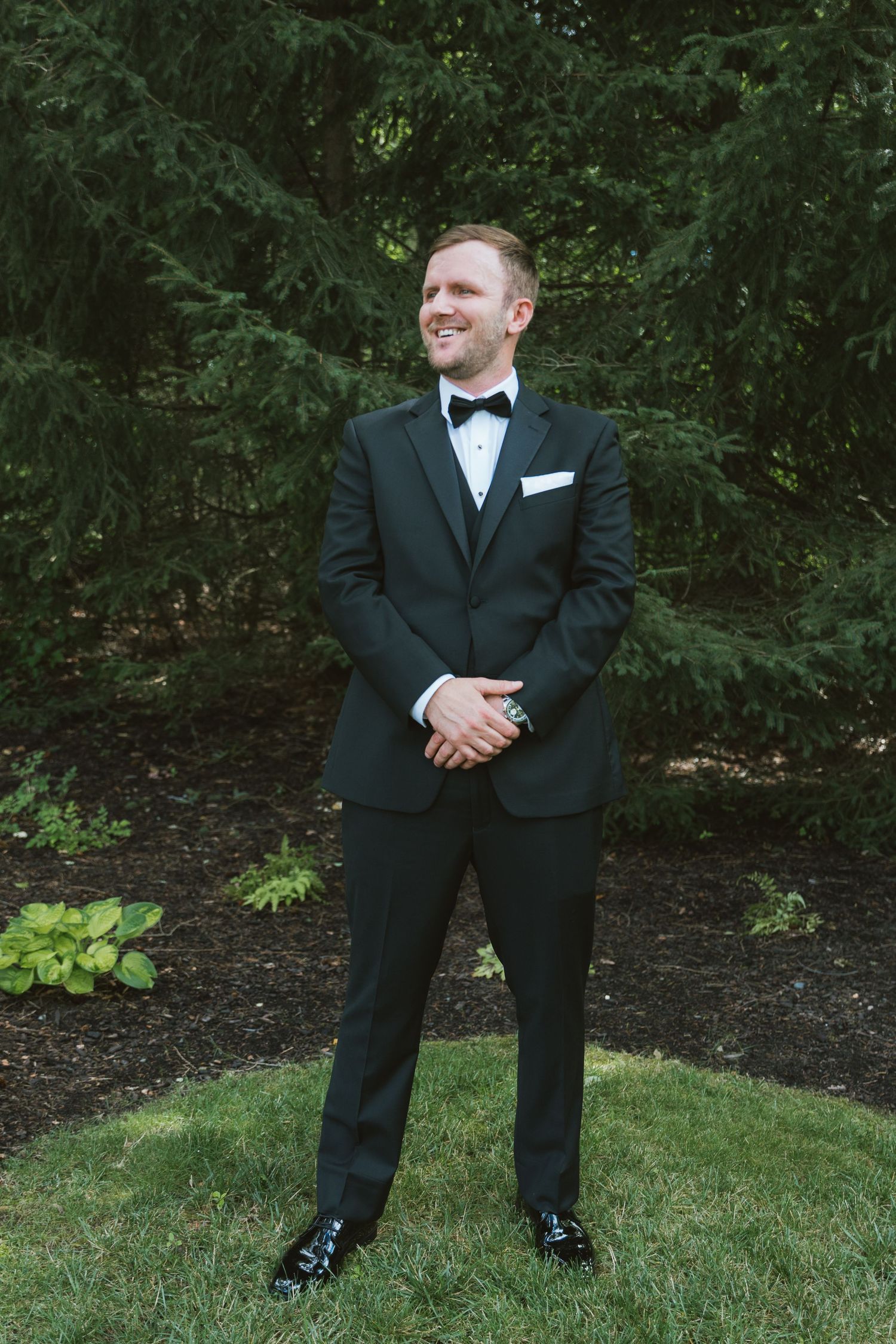 A smiling groom in a black tuxedo with bow tie and white pocket square stands outdoors among lush green trees.