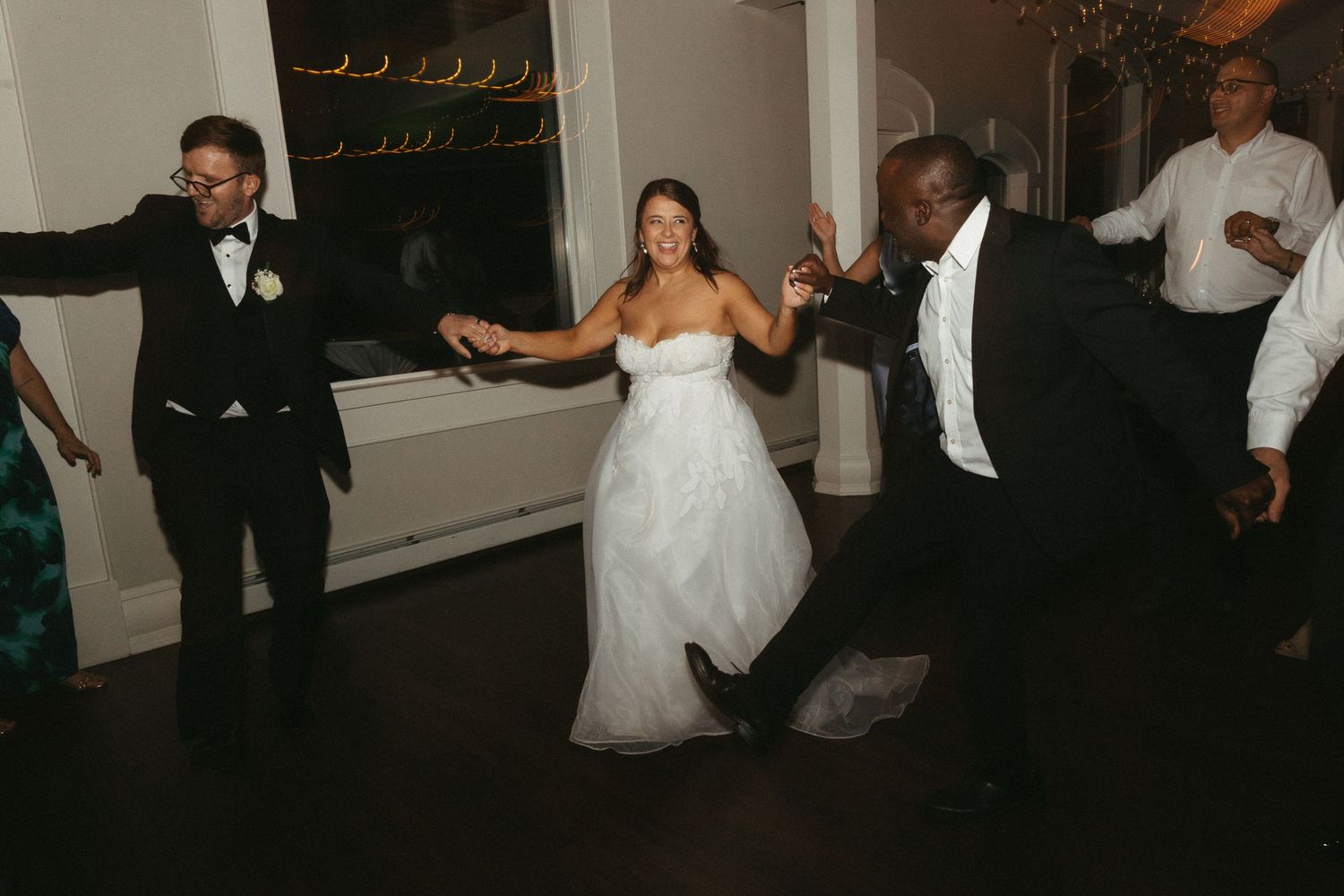 Bride in white gown dances joyfully with guests in formal attire at a wedding reception celebration.