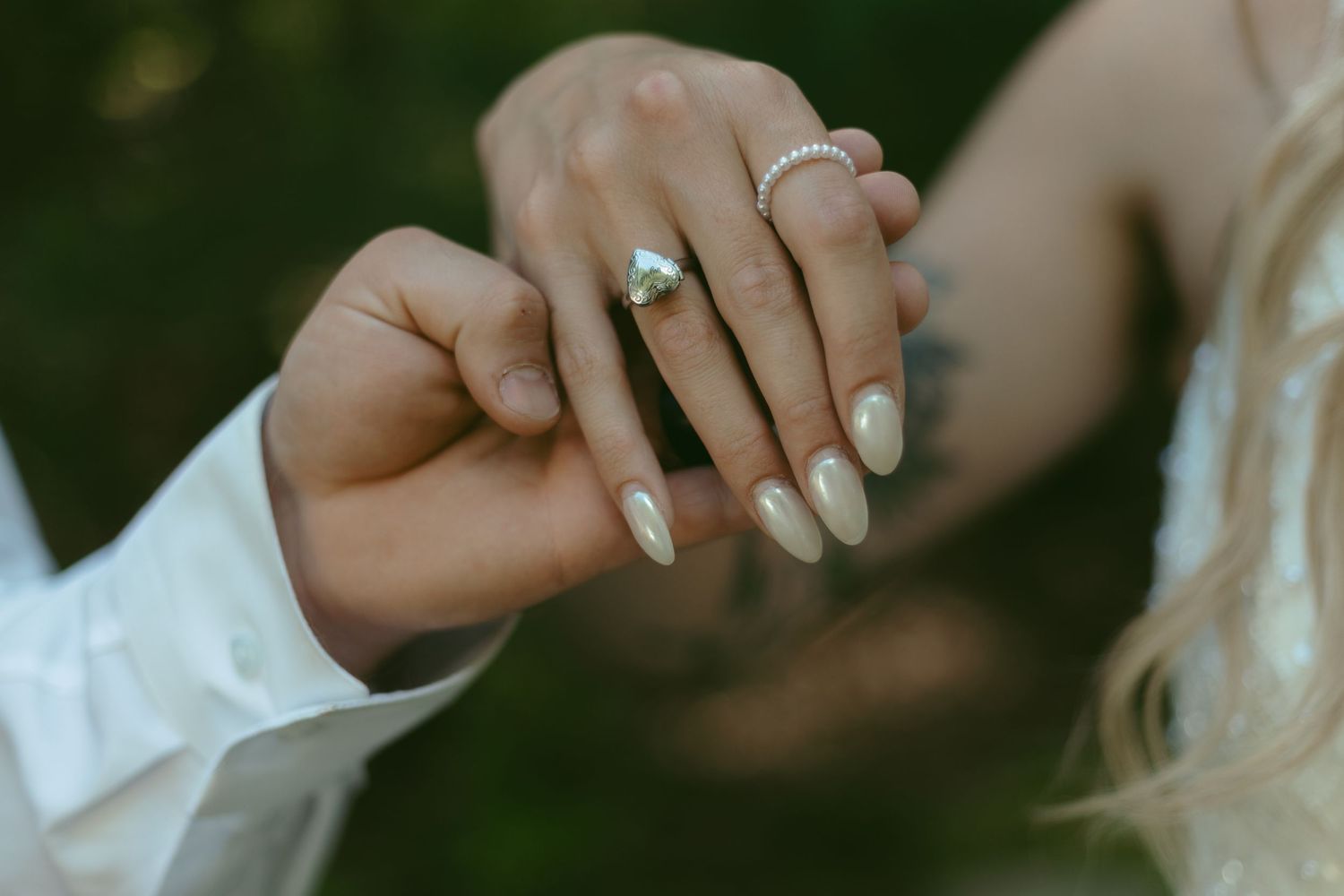 Close-up of bride's hand with diamond engagement ring and pearl ring, holding groom's hand outdoors.