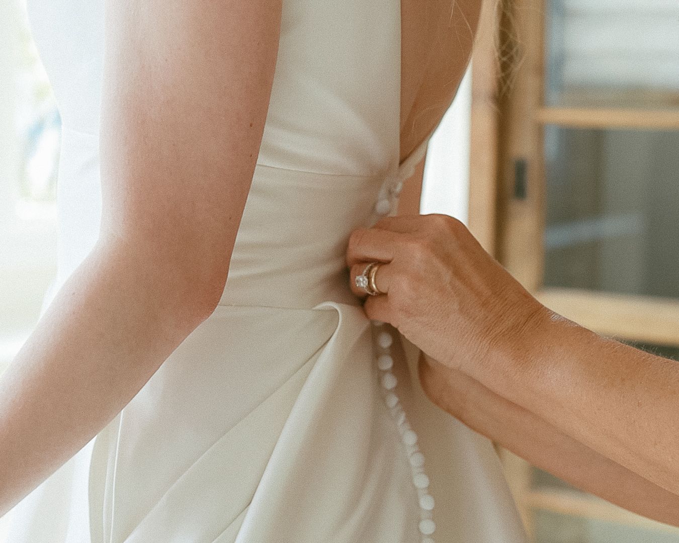 Candid photo of a mother with gold rings buttoning the back of her daughter’s modern crepe wedding gown in Lehigh Valley, PA.