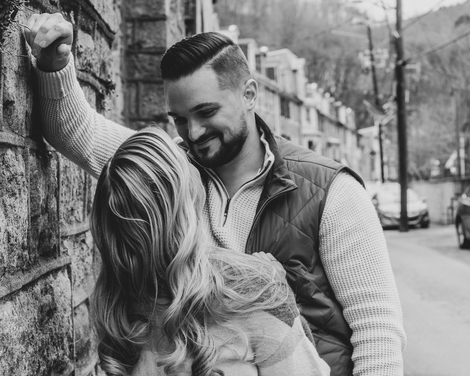 Black and white photo of couple embracing against brick wall on a city street.