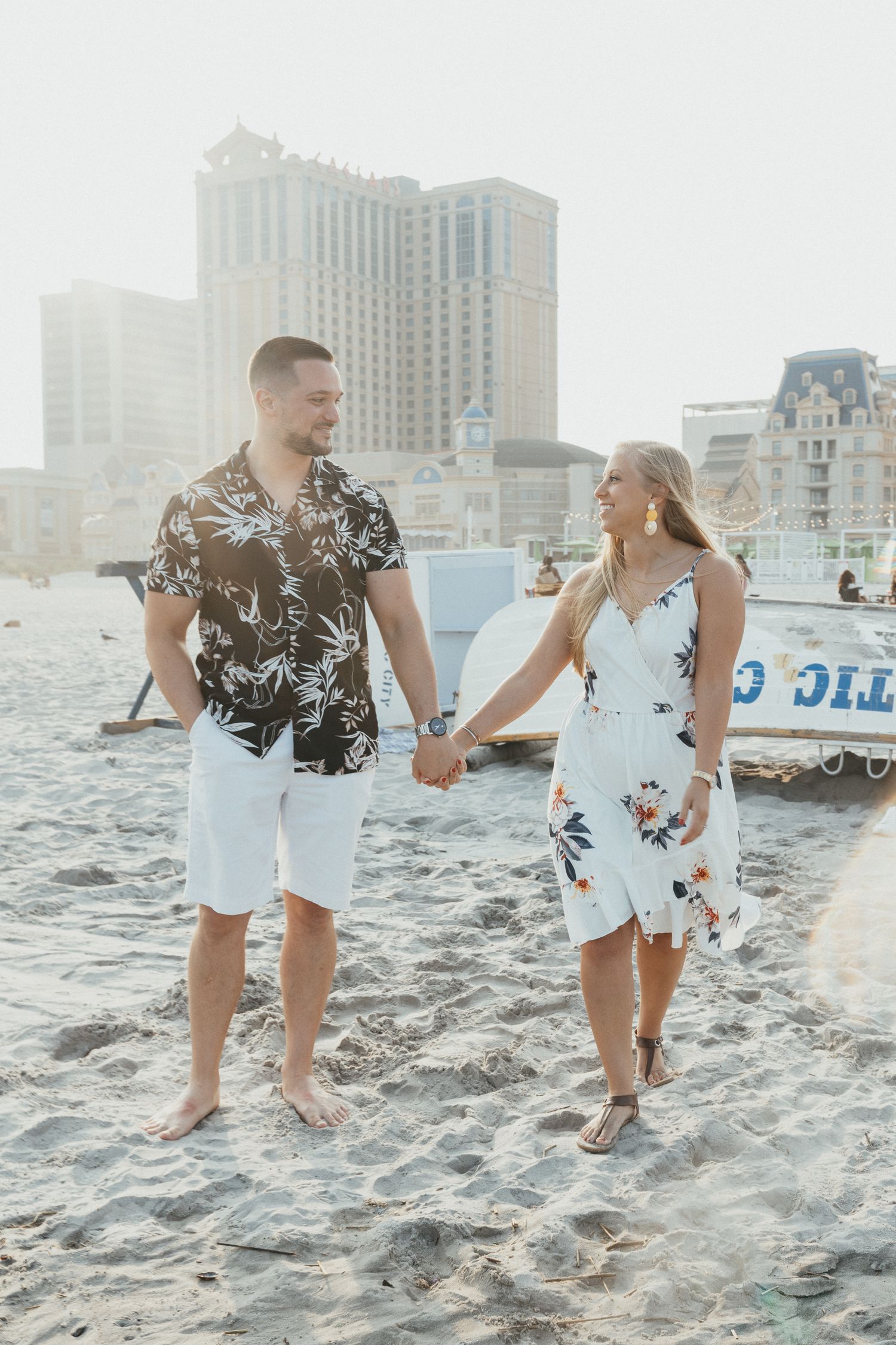 Couple holding hands on beach with Atlantic City skyline in background, man in floral shirt, woman in white dress.