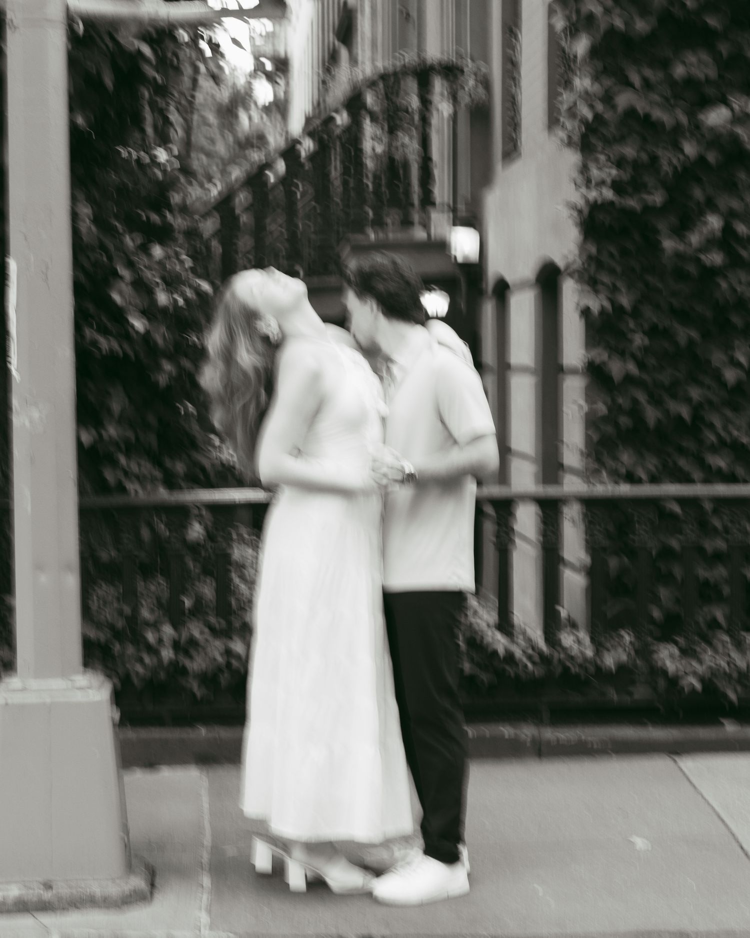 Black and white photo of a couple embracing on a city sidewalk, surrounded by ivy-covered walls.
