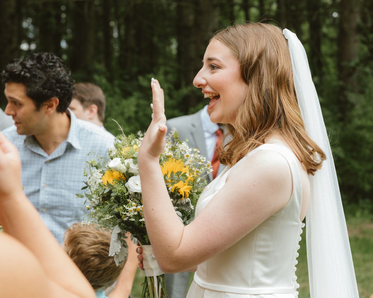 Smiling bride in white dress holding sunflower bouquet waving at outdoor wedding ceremony.