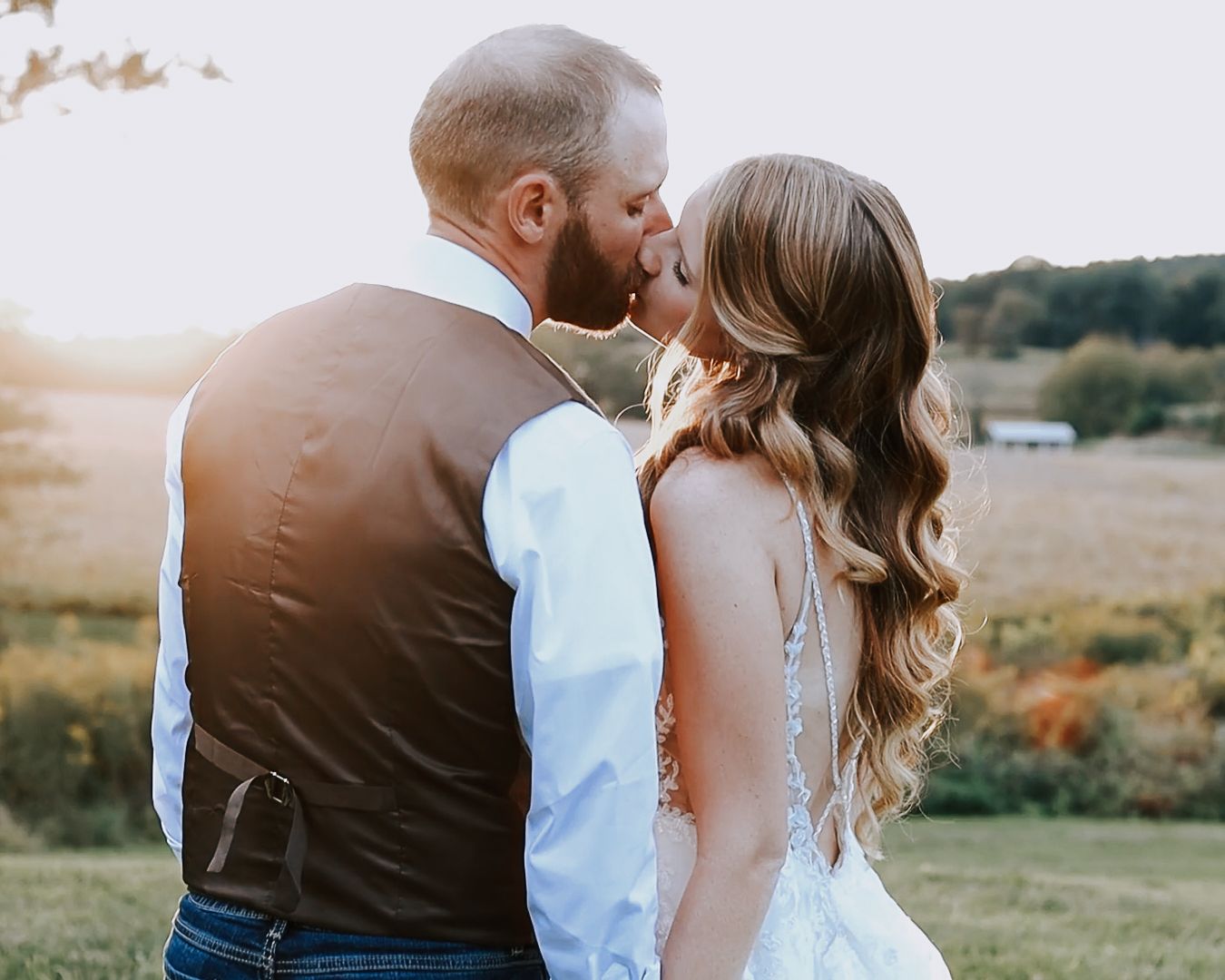 A bride and groom share a romantic kiss outdoors at sunset during their rustic countryside wedding ceremony.