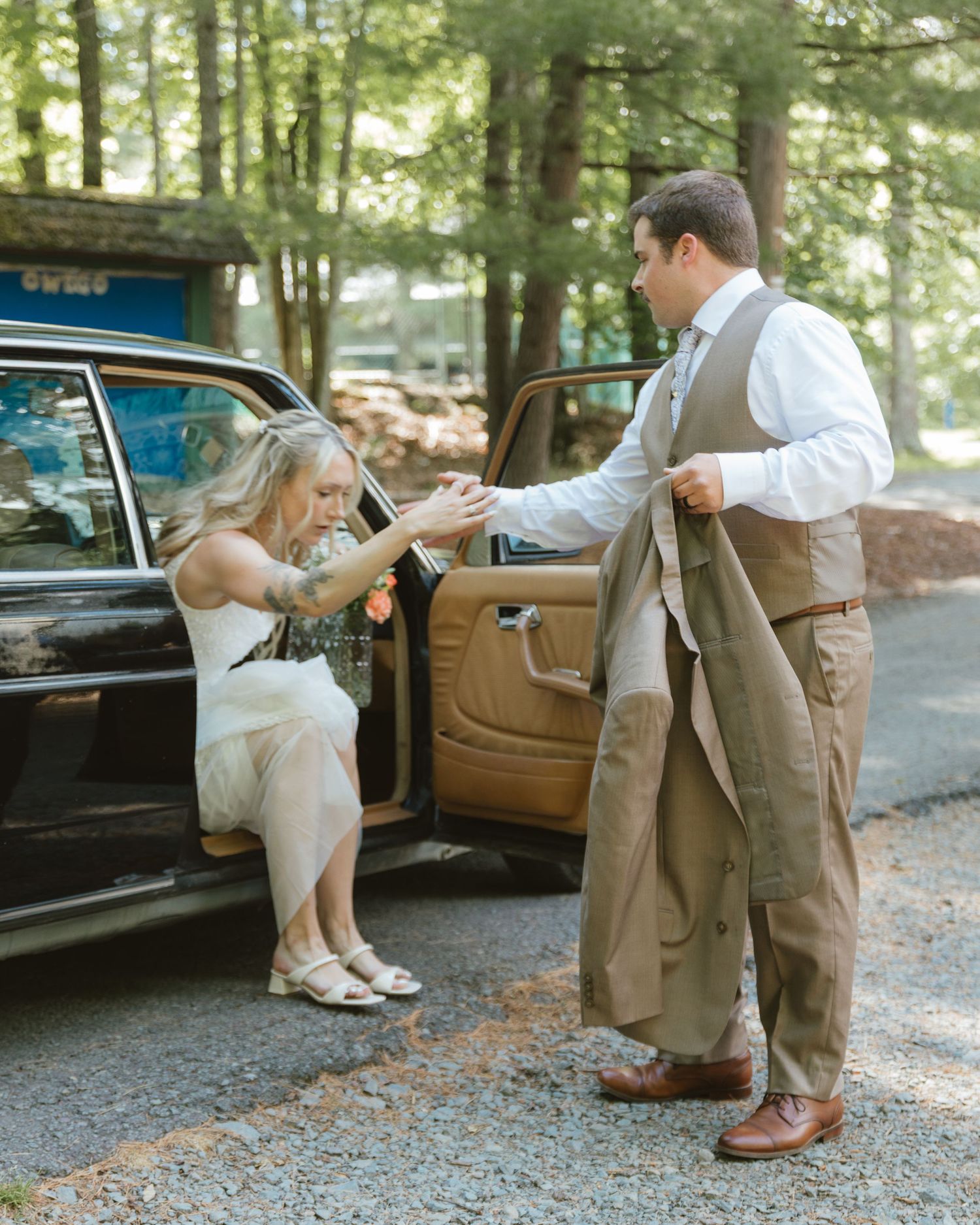 A groom helps a bride exit a vintage black car outdoors near trees at a rustic wedding venue.