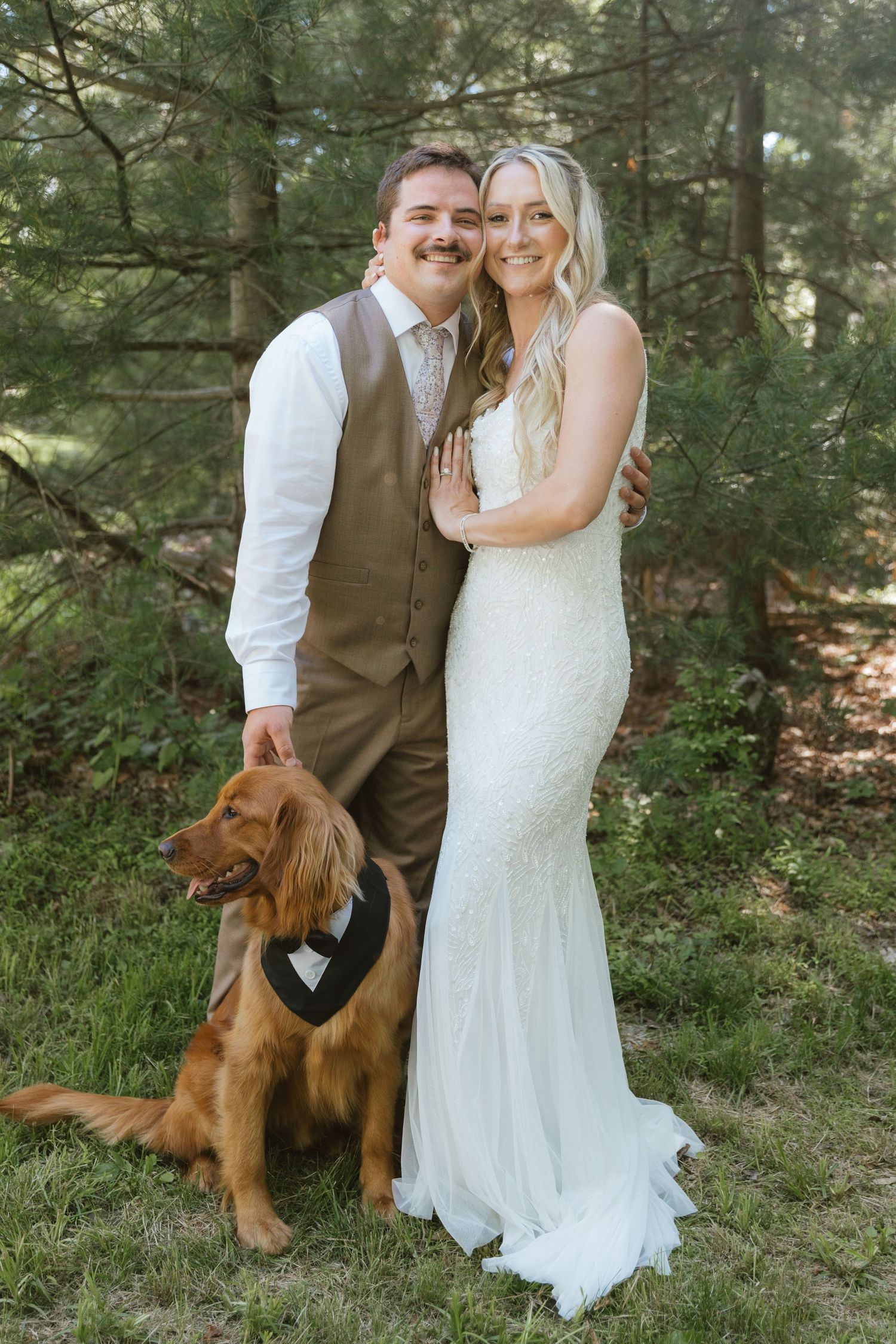 Bride in white gown and groom in tan vest pose with golden retriever wearing bow tie in forest setting.