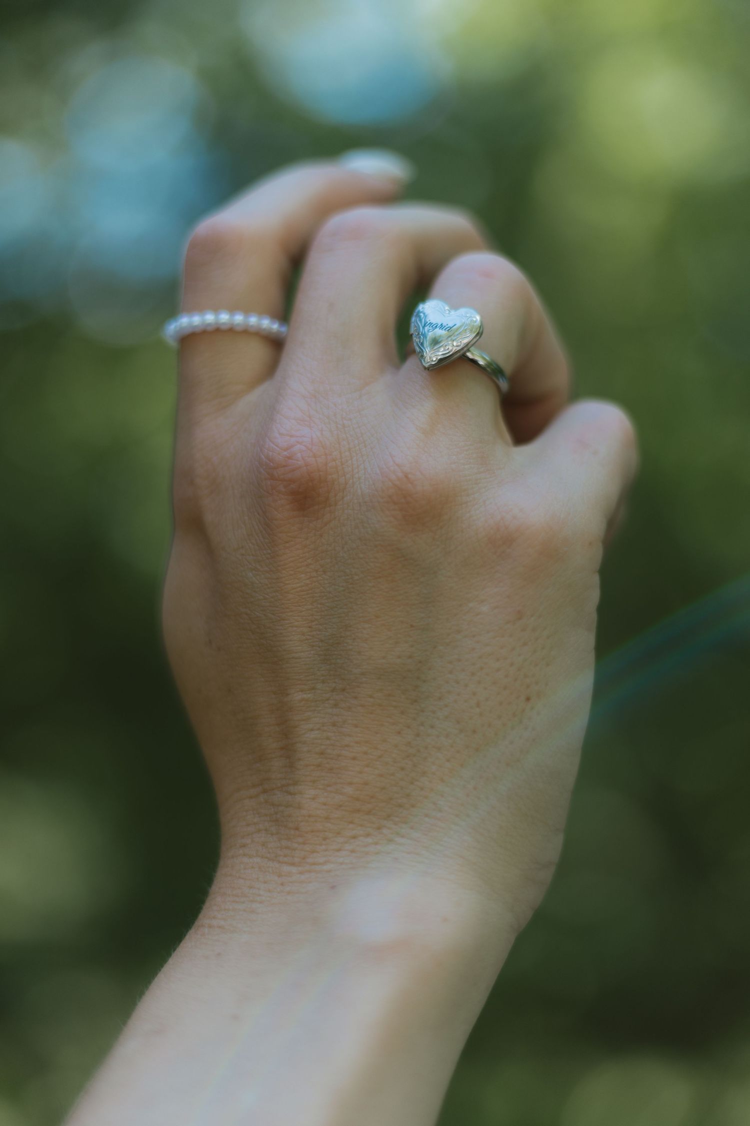 Close-up of a hand wearing a heart-shaped diamond engagement ring and delicate pearl band outdoors.