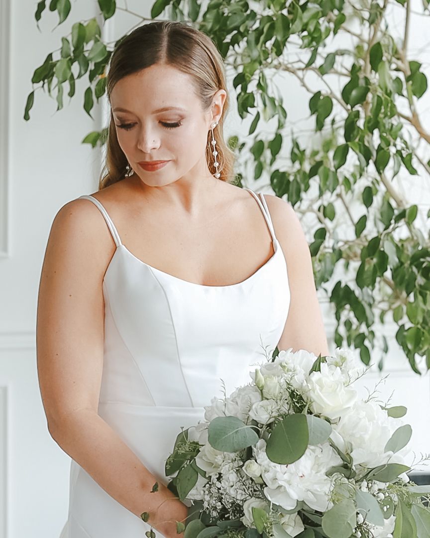 Elegant bride in simple white slip gown holds lush white and green bouquet near leafy backdrop.