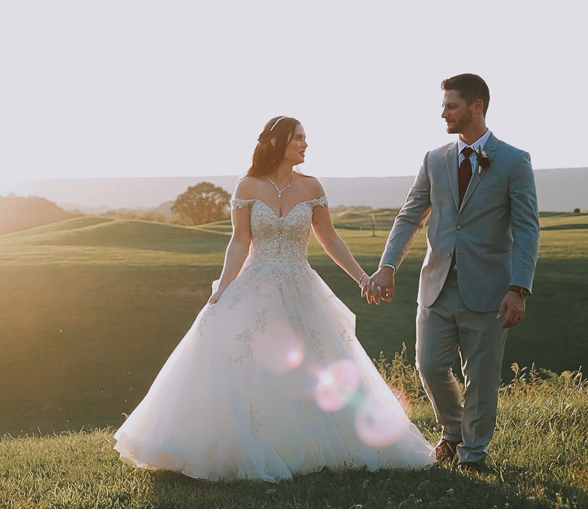 Bride and groom holding hands at golden hour on a green hillside, bride in off-shoulder ballgown.