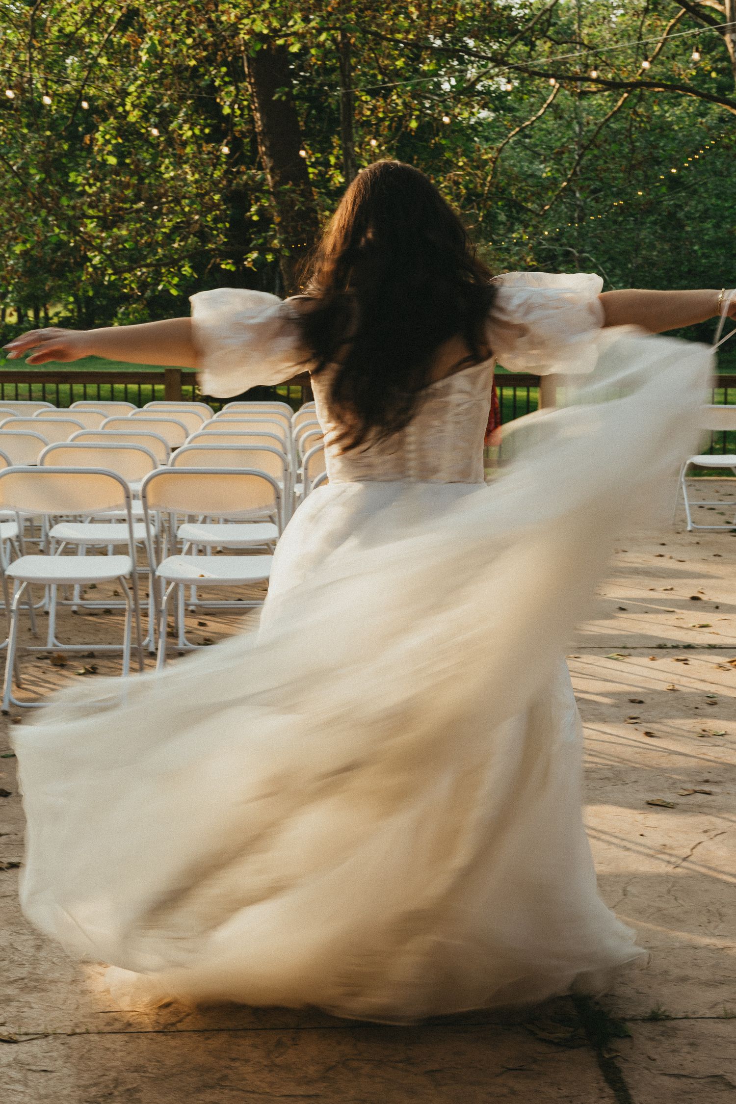 Bride in flowing white tulle gown twirling with arms outstretched near ceremony chairs in garden.