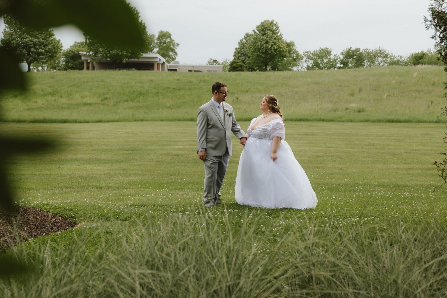 Bride in white tulle gown and groom in gray suit hold hands walking through lush green field at outdoor wedding venue.