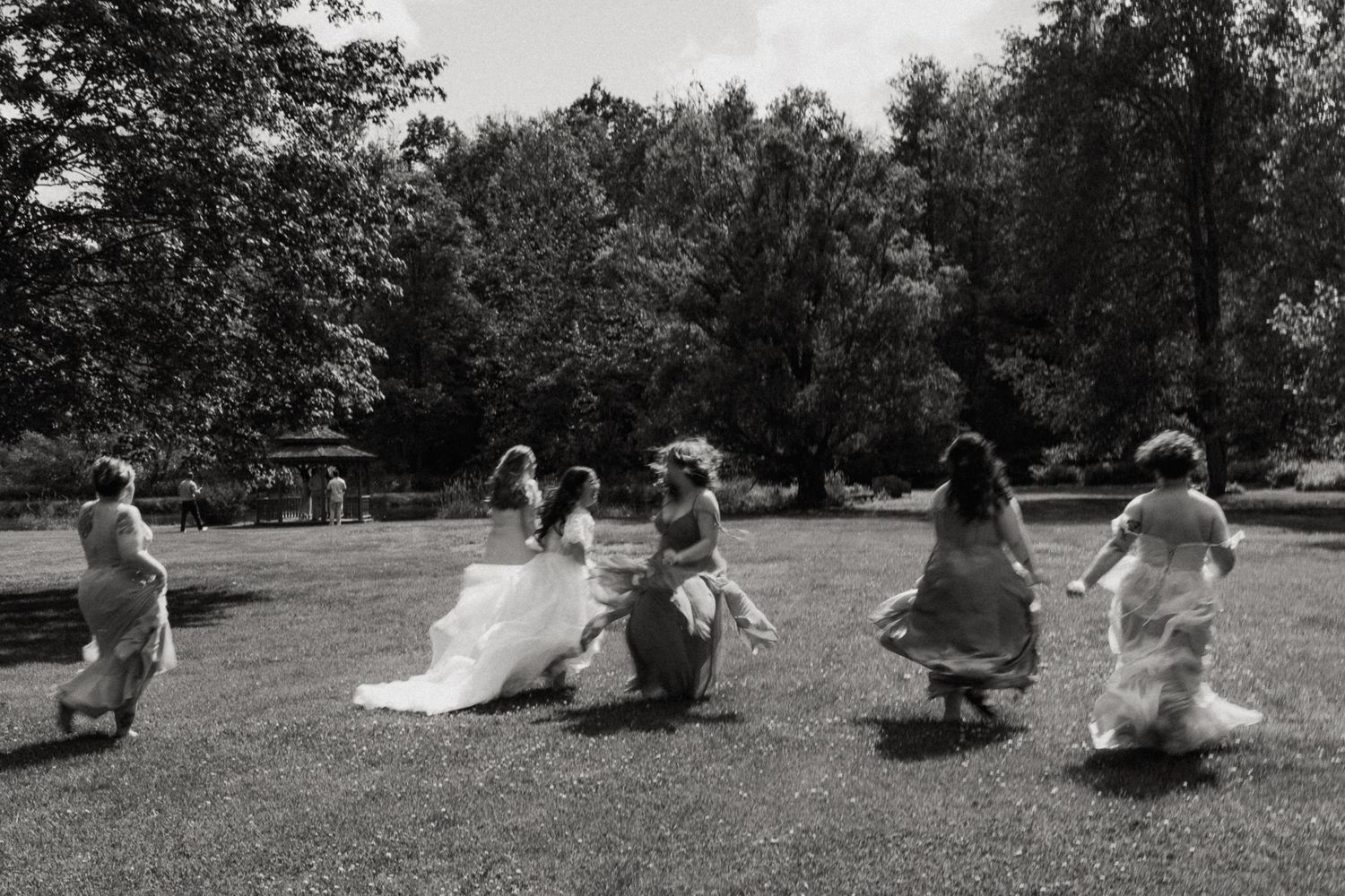 Black and white photo of bride and bridesmaids running joyfully across garden lawn.