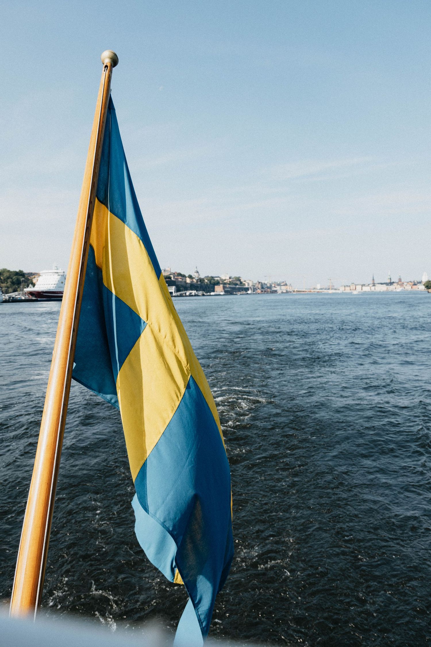 Swedish flag waving from a boat stern with Stockholm cityscape across blue waters.