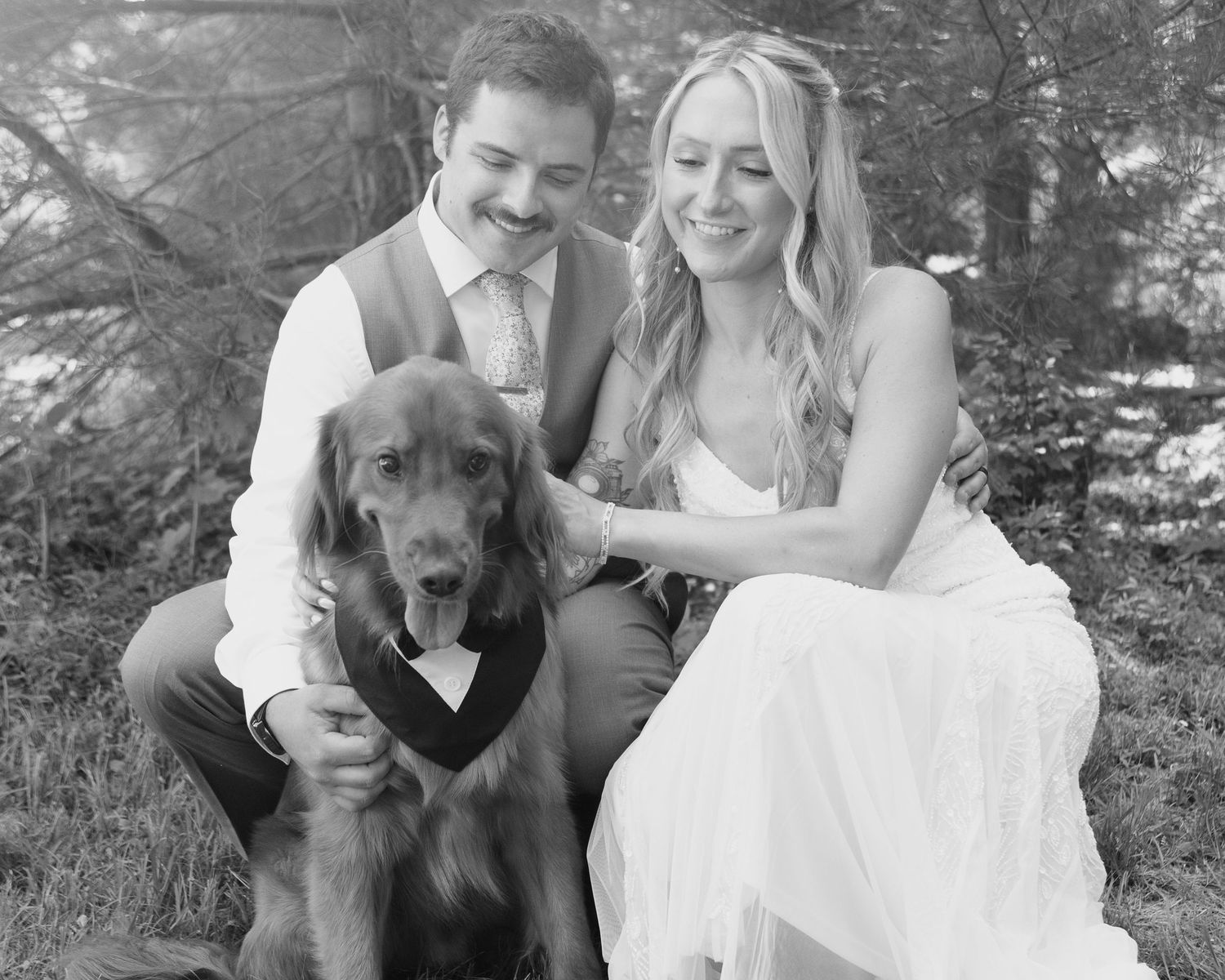 Black and white wedding photo of smiling couple in formal attire posing with their dog wearing a bow tie outdoors.