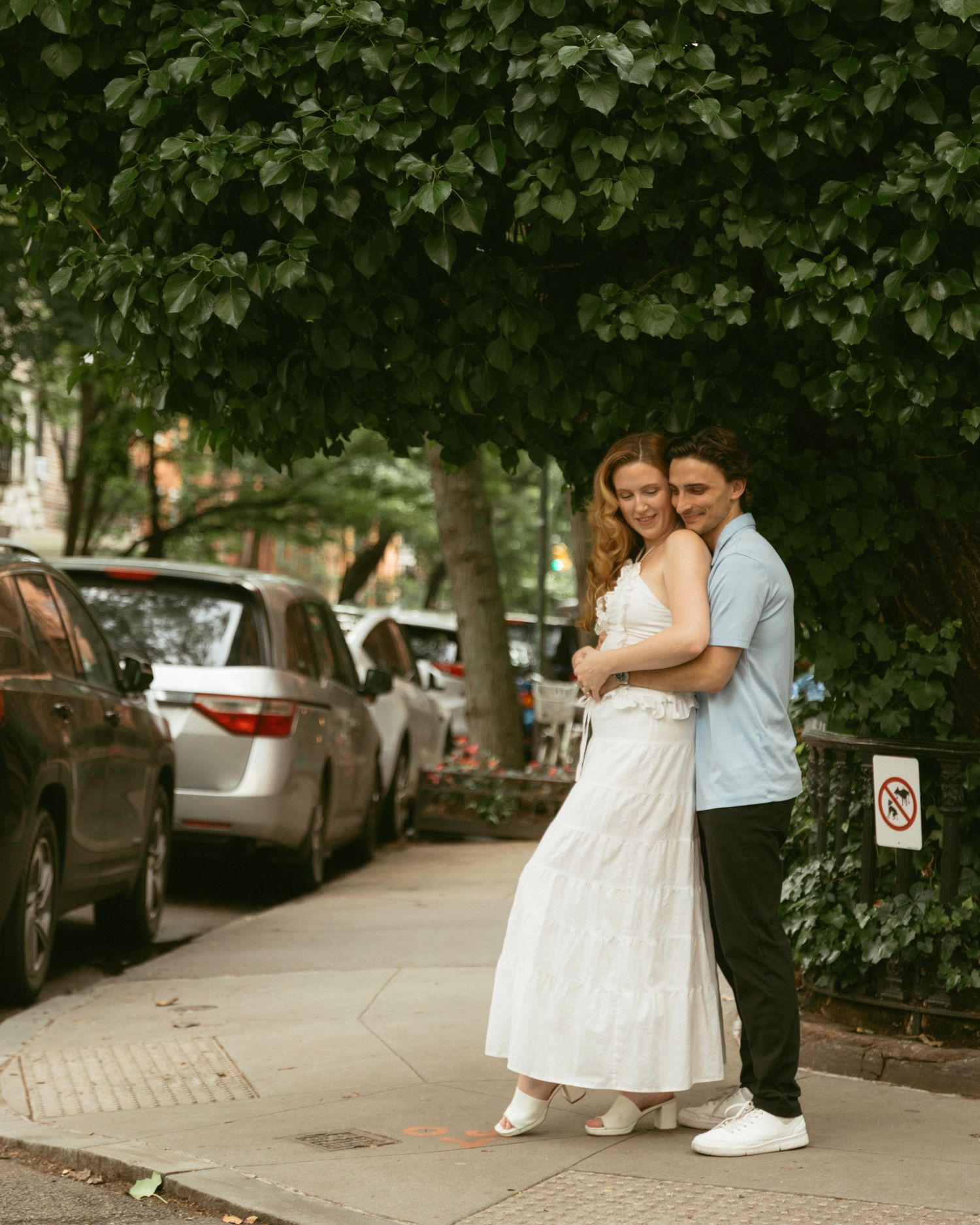 Couple embracing on a tree-lined urban sidewalk, woman in white dress, man in blue shirt.