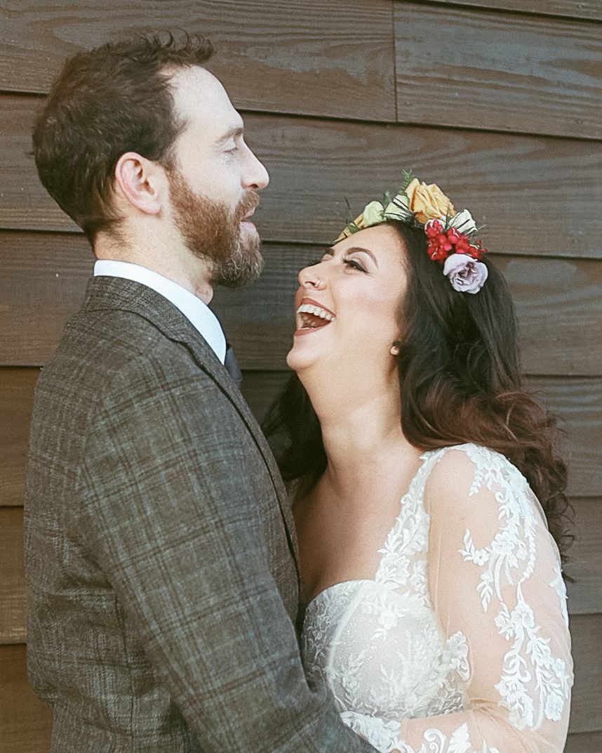 Candid portrait of a joyful couple in front of a rustic wood wall, featuring a boho floral crown from an Allentown wedding.