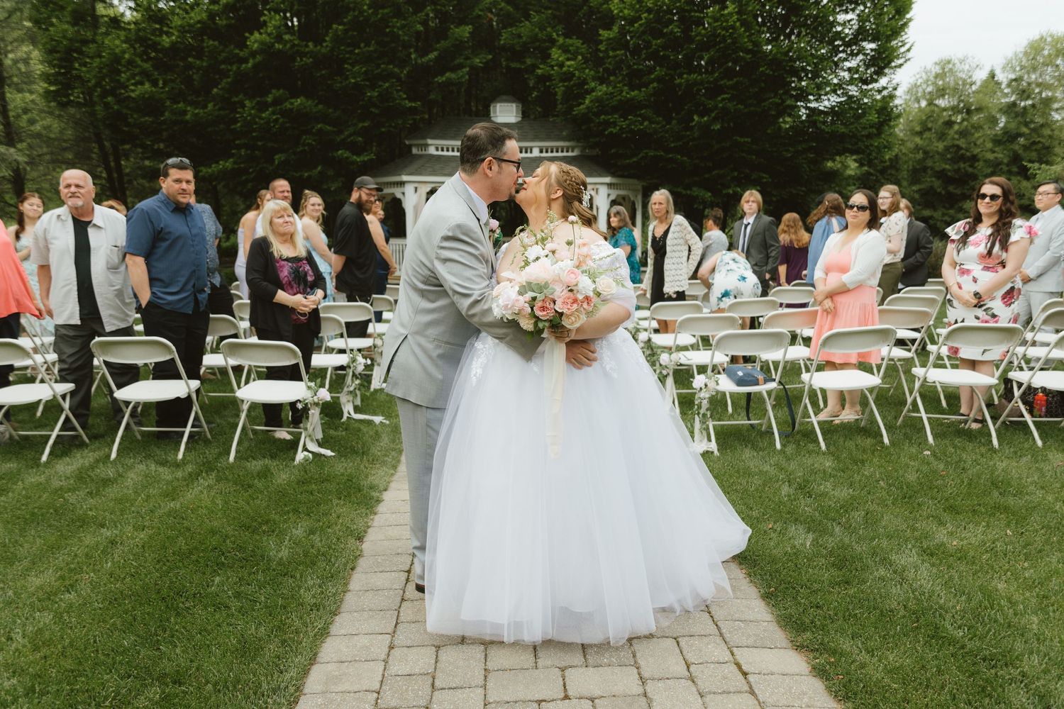 Bride and groom share a kiss at outdoor wedding ceremony on brick path surrounded by guests seated in white chairs.