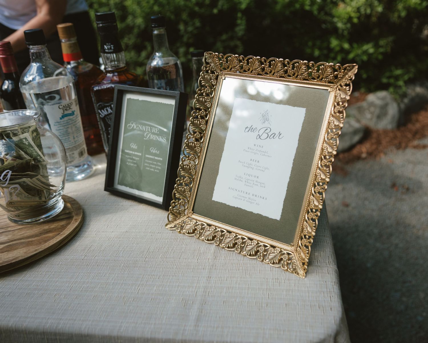 Gold ornate framed bar menu sign displayed on a draped table at an outdoor wedding reception.