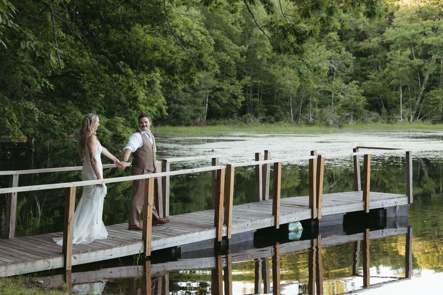 Bride and groom holding hands on a wooden dock surrounded by lush green trees and a serene pond.