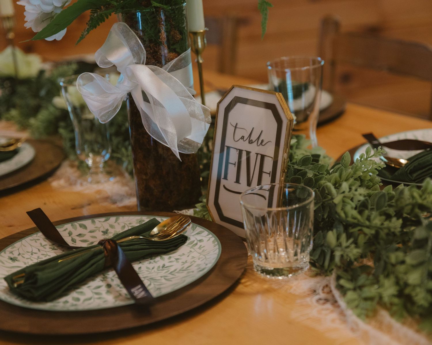 Elegant wedding table setting with greenery, gold cutlery, and a Table Five sign at a rustic reception.