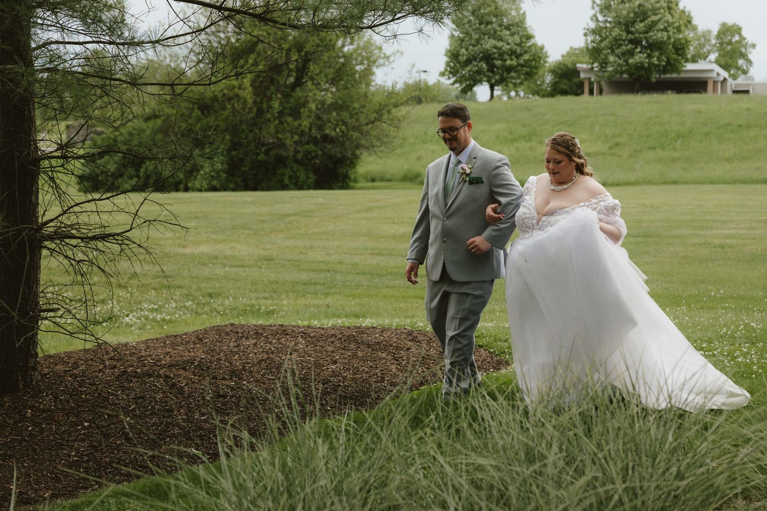 Bride in white gown and groom in sage suit walk together across lush green lawn during outdoor wedding.