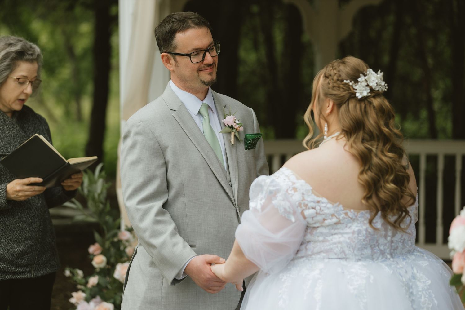 Groom in gray suit holds bride's hands during outdoor wedding ceremony while officiant reads from book.