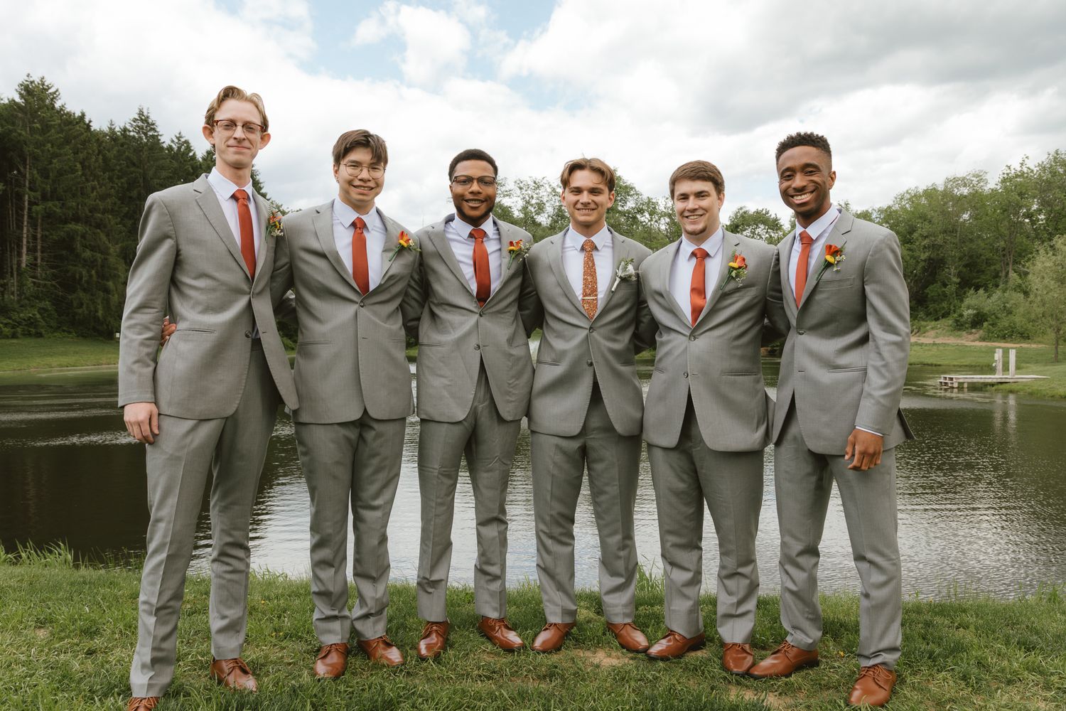 Six groomsmen in matching gray suits with orange ties pose outdoors near a pond on a wedding day.