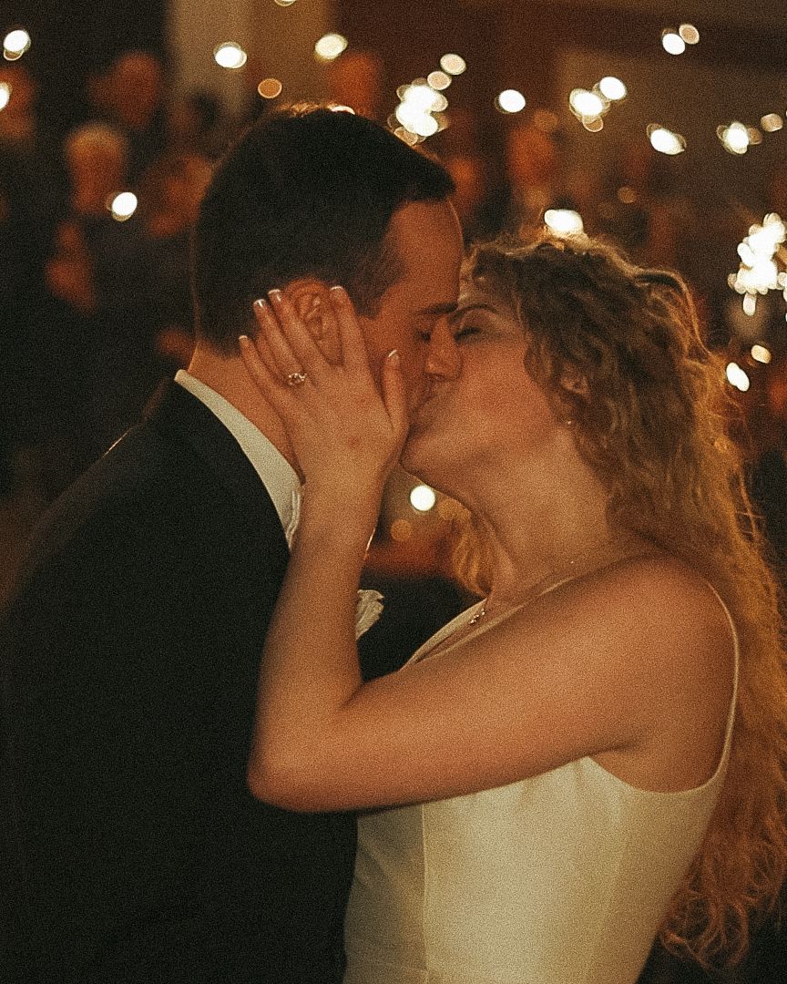 Bride and groom share romantic first kiss on dance floor surrounded by warm glowing bokeh lights.