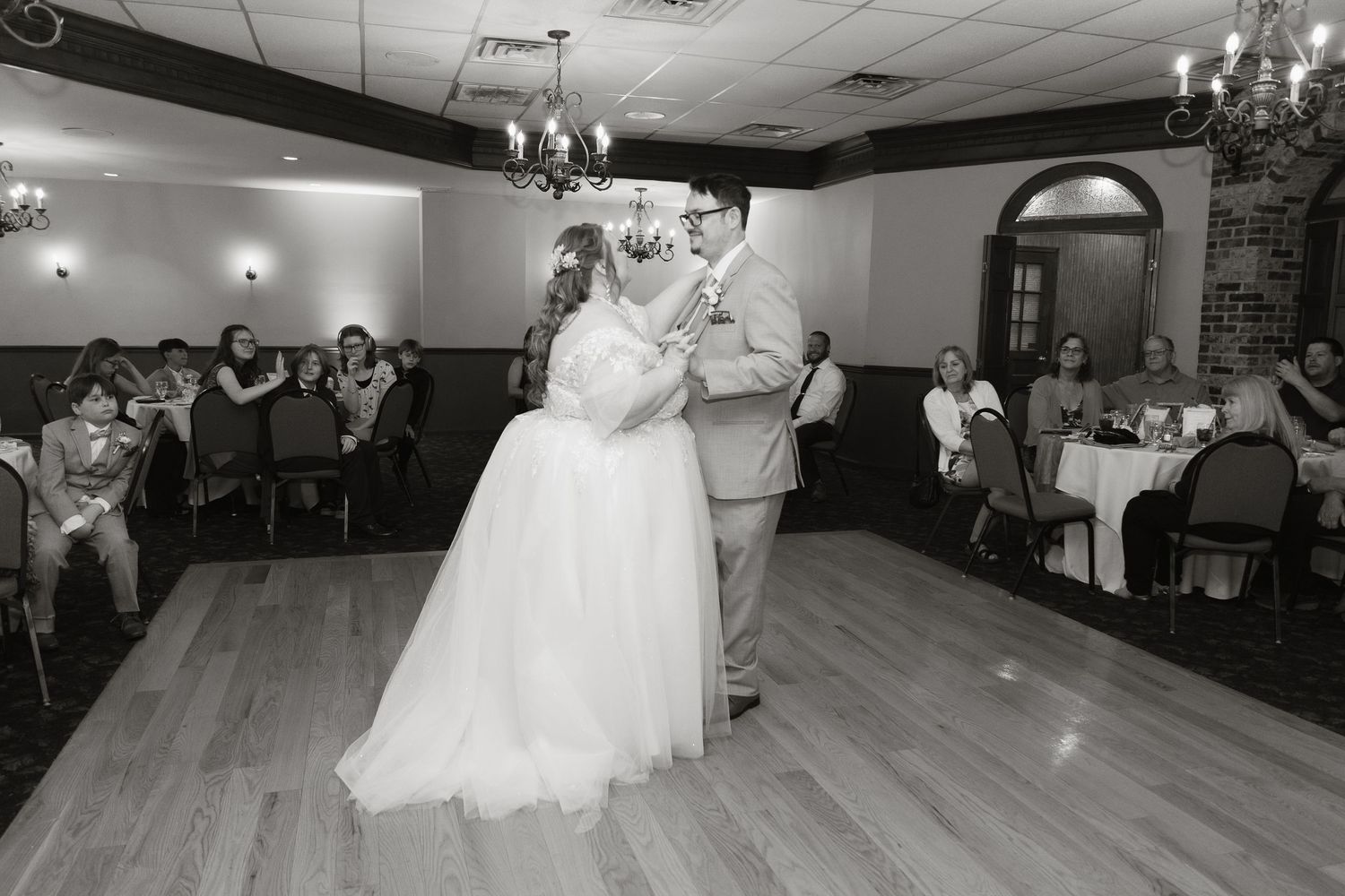 Black and white photo of a bride and groom sharing their first dance at a wedding reception with guests watching.