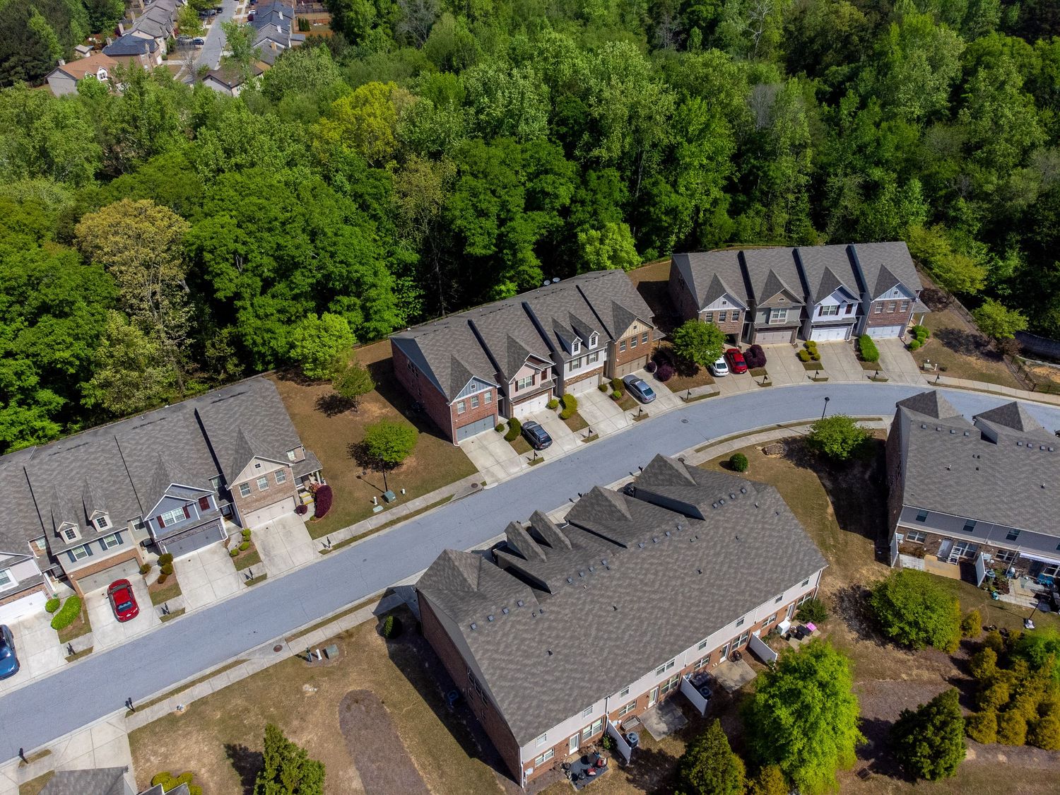 Aerial view of suburban houses with grey roofs along a curved residential street surrounded by lush green trees.