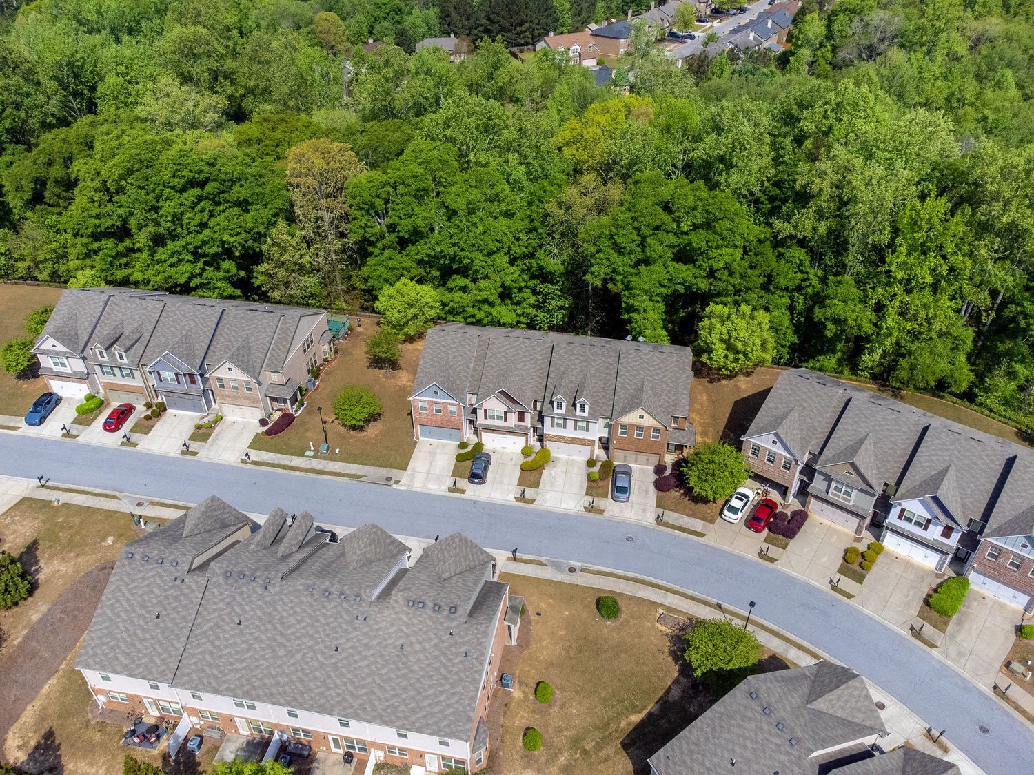 Aerial view of modern townhouses with gray roofs arranged along a curved suburban street surrounded by green trees. real estate photography
