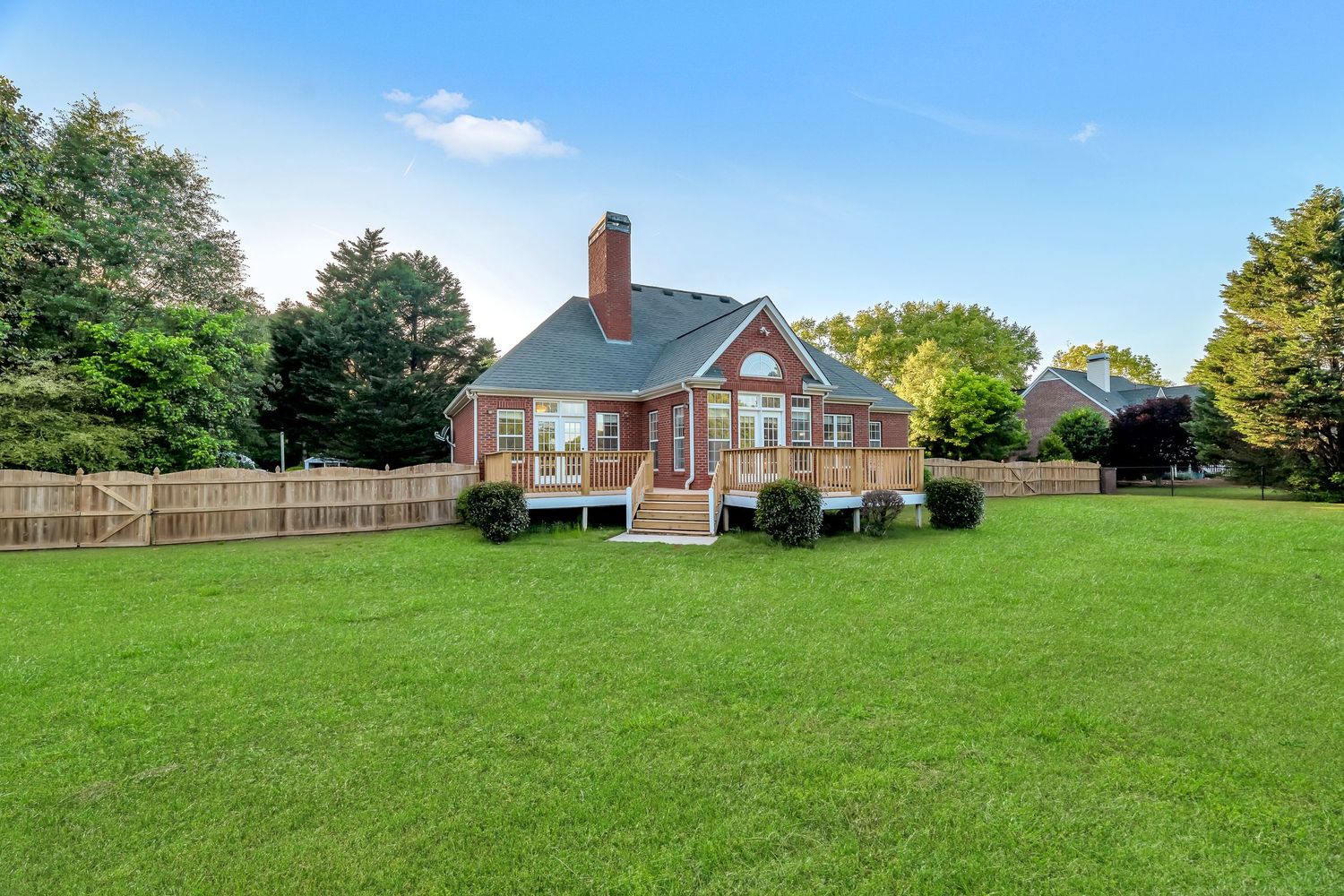 Large green grassy field with a red brick house and trees visible in the background on a sunny day.