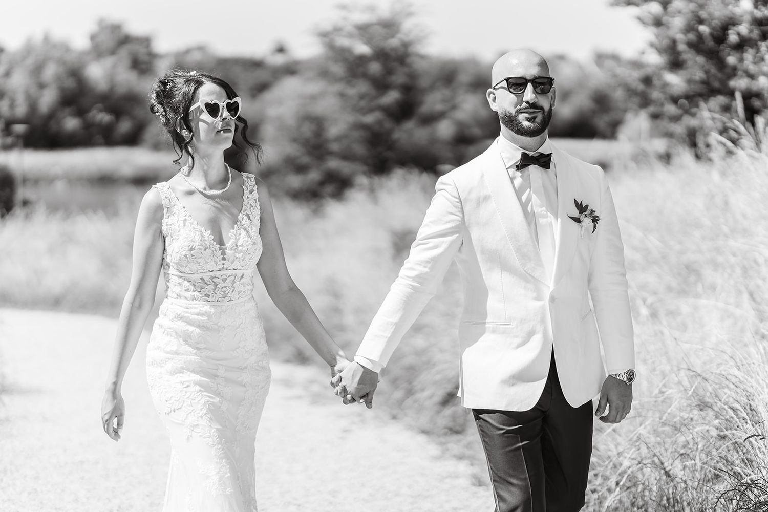 A black and white wedding photo of a couple walking hand in hand through a sunny meadow.