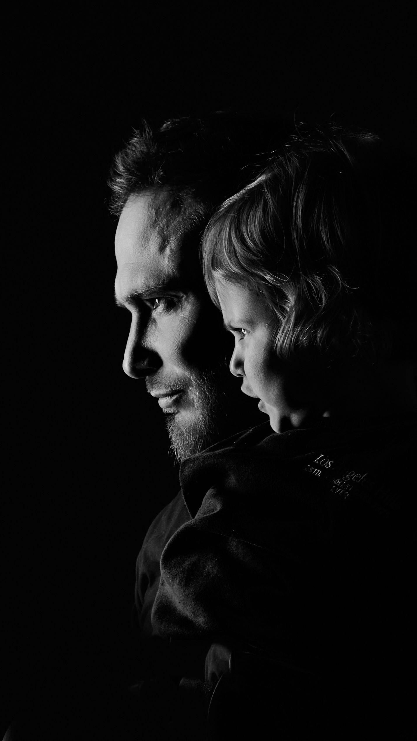 Black and white portrait of a smiling father holding his young child, both in profile against a dark background.